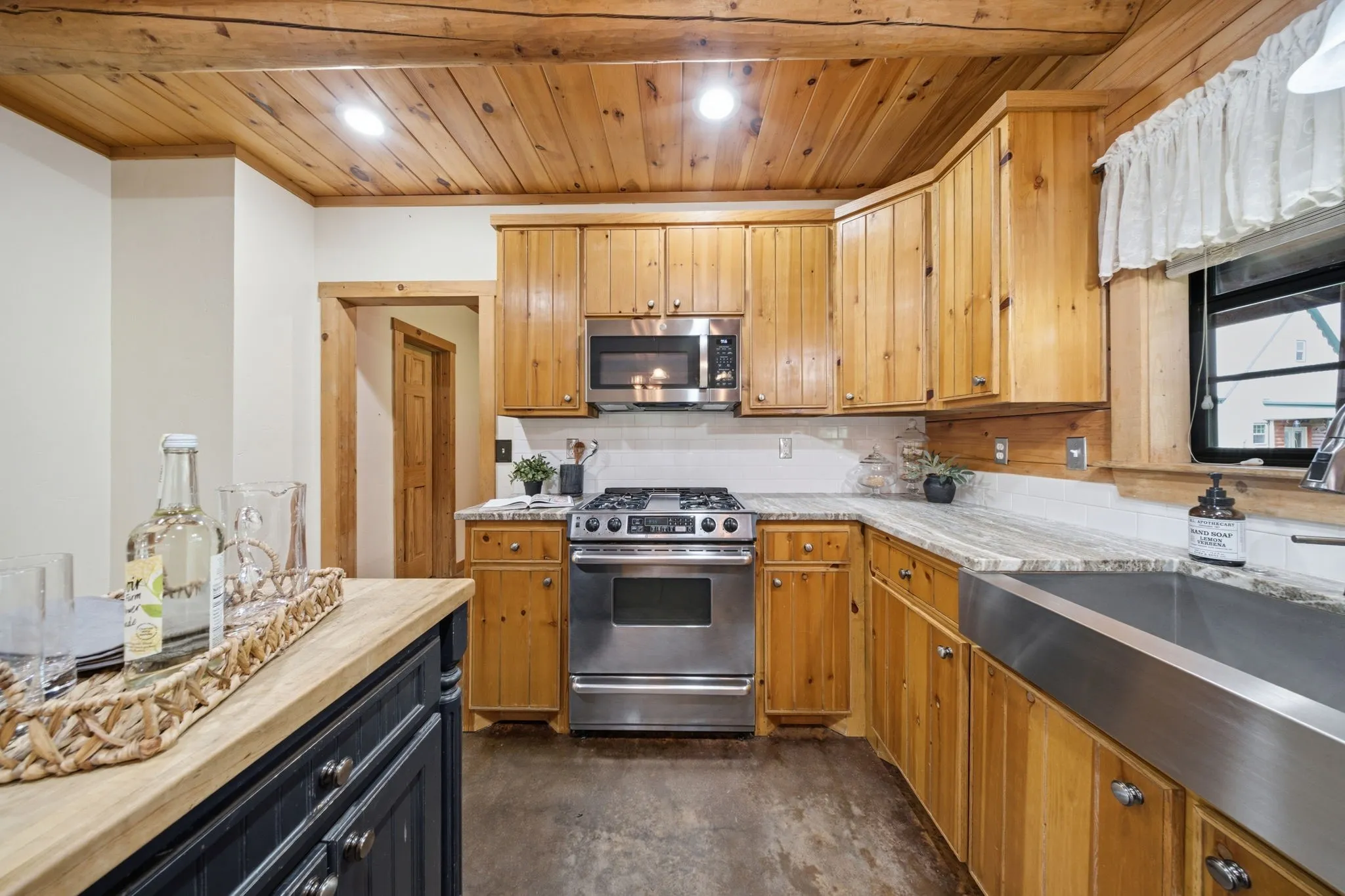 Wood-paneled ceiling, stainless steel appliances, farmhouse-style sink, light-toned countertops, and natural wood cabinetry