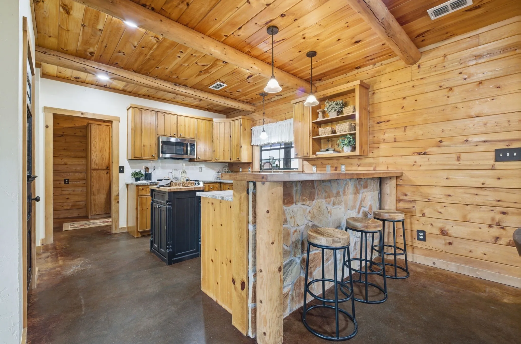 Wood-paneled walls and ceiling with exposed log beams, stone-faced breakfast bar, and stainless steel microwave