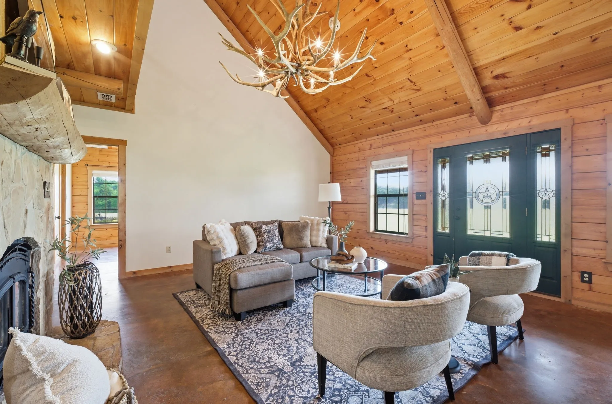 Vaulted wood-paneled ceiling, stone fireplace with a wood mantel, stained concrete flooring, and an antler chandelier
