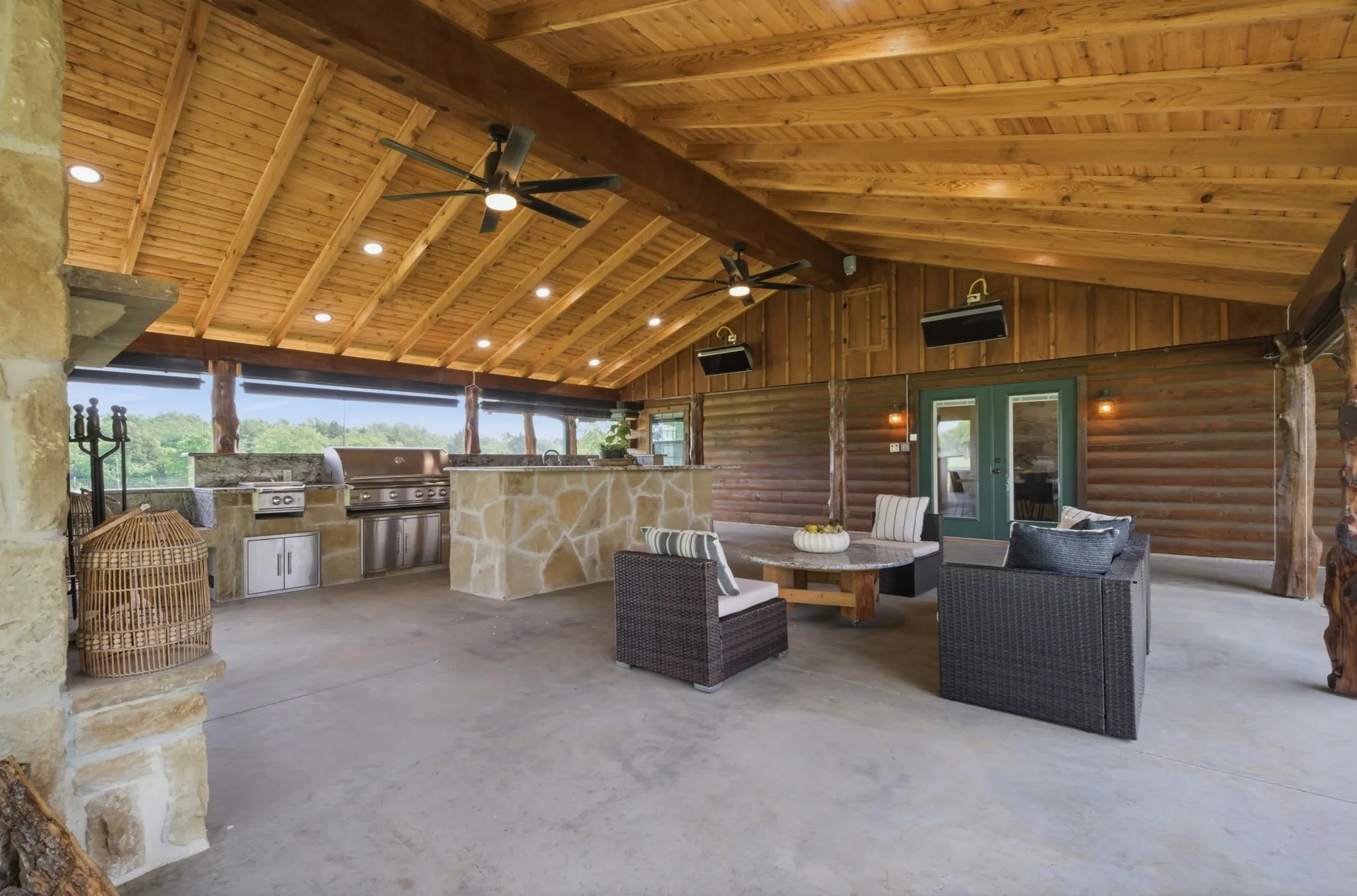 Expansive covered patio featuring a vaulted wood ceiling, built-in outdoor kitchen with stainless steel appliances, stone-clad bar, and concrete flooring