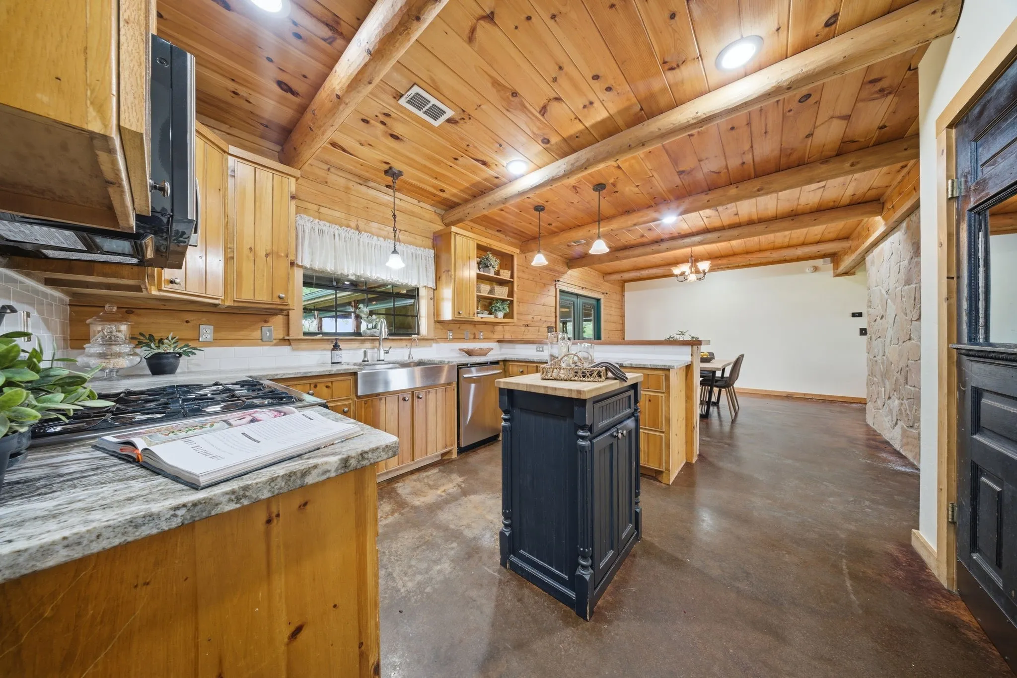 Wood-paneled ceilings with exposed beams, light wood cabinetry, stone-finish countertops, stainless steel appliances, and a central island with a contrasting dark finish