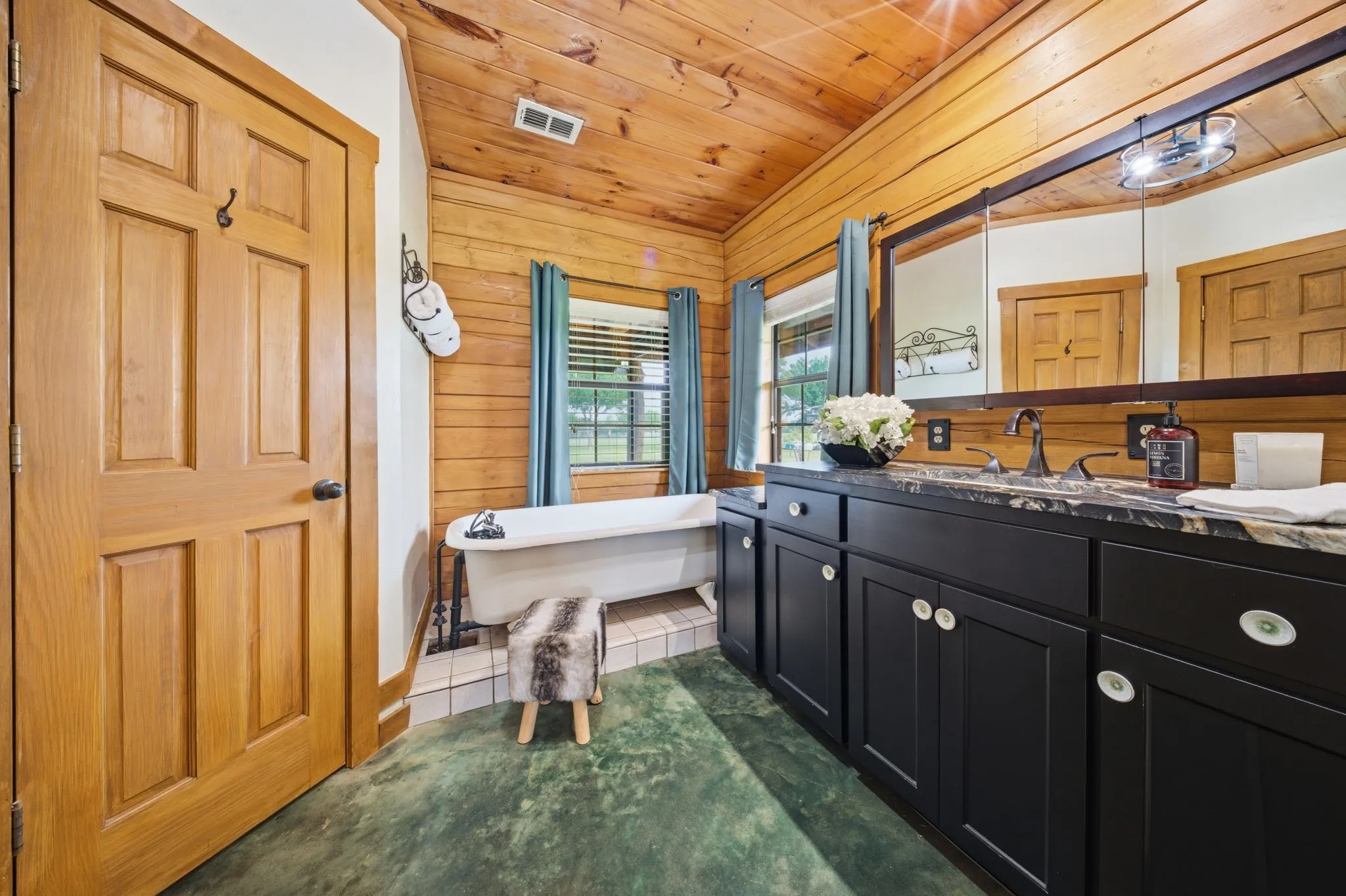 Wood-paneled walls and ceiling, classic clawfoot tub, double vanity with dark cabinetry, integrated lighting over vanity, and a window with horizontal blinds