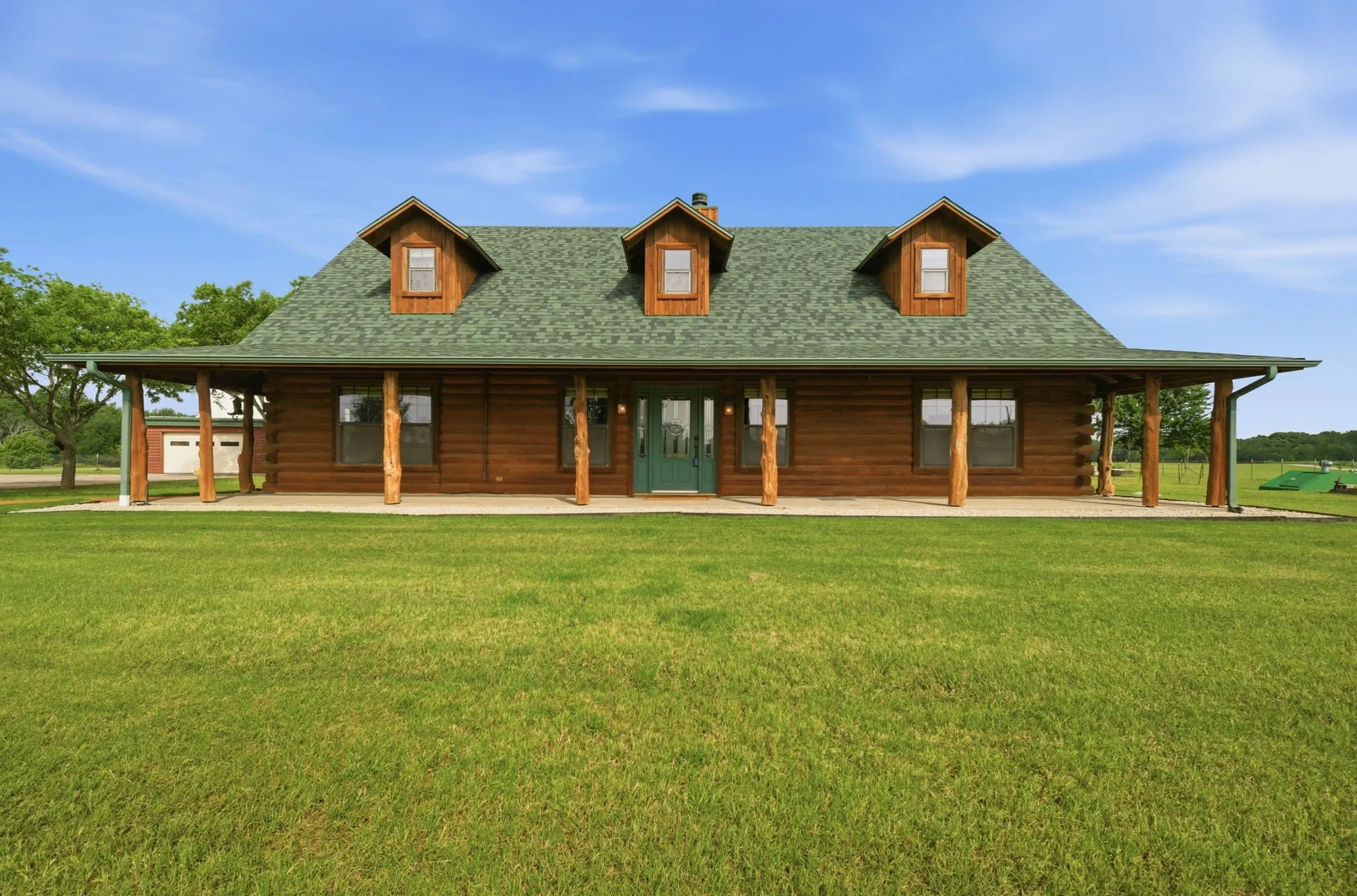 Log cabin exterior with an expansive covered porch, natural wood support columns, three dormer windows, and a green shingle roof