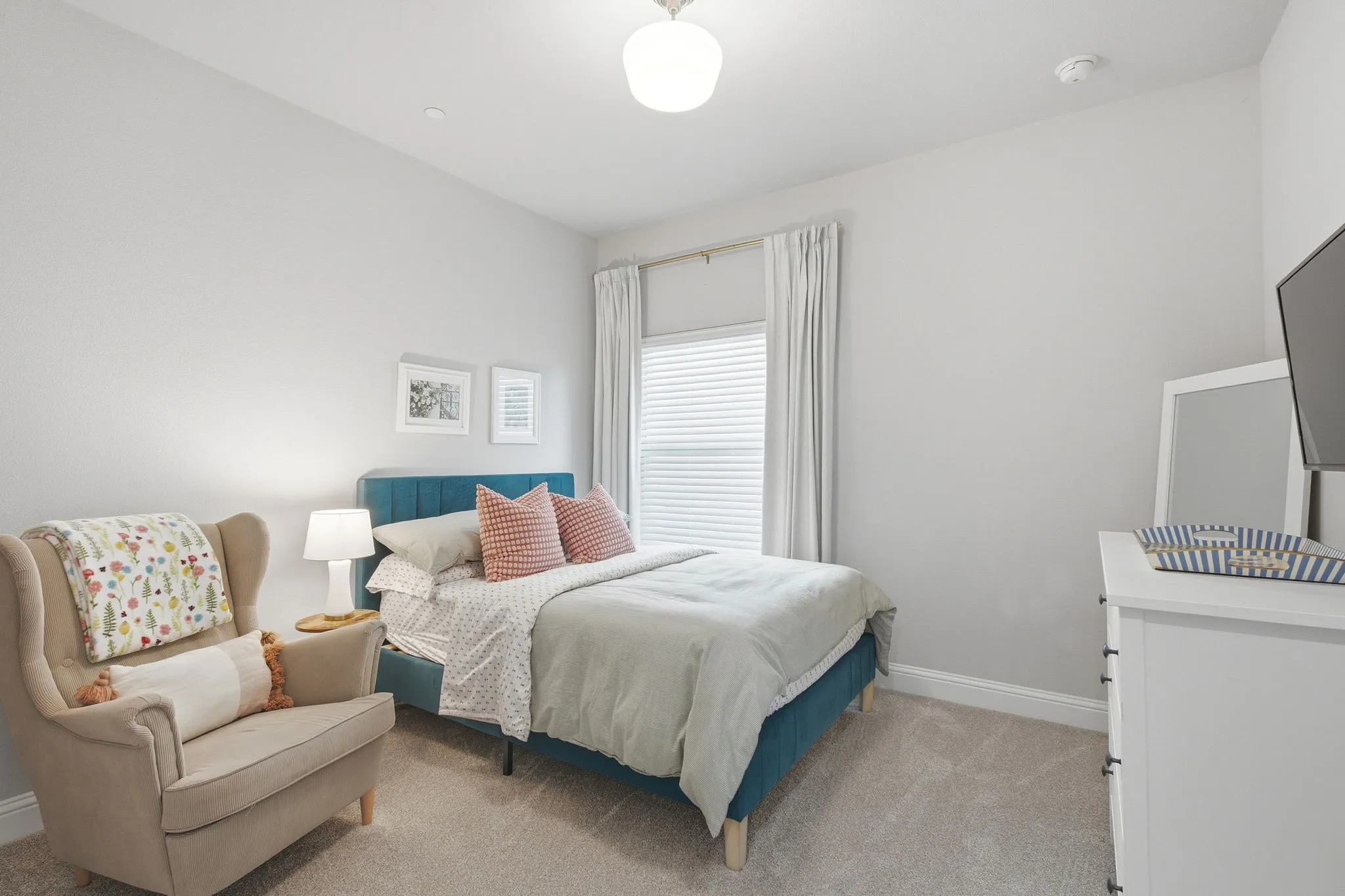 Carpeted flooring, window with blinds and curtains, light neutral wall paint, overhead light fixture, and a built-in smoke detector