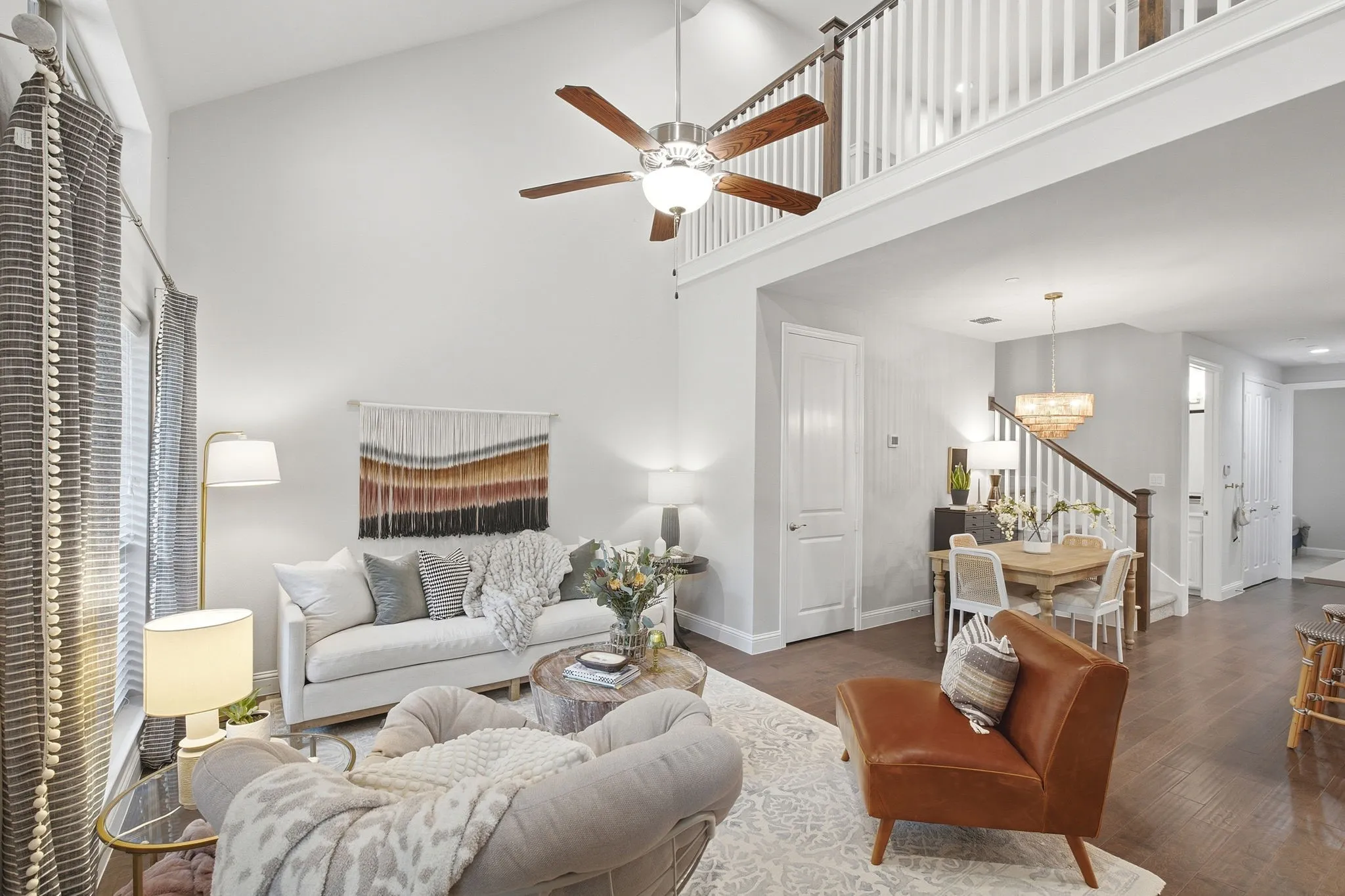 Vaulted ceiling with ceiling fan, wood-finish flooring, white painted interior walls, second-story interior balcony with white spindle railing, and a dining area with a chandelier