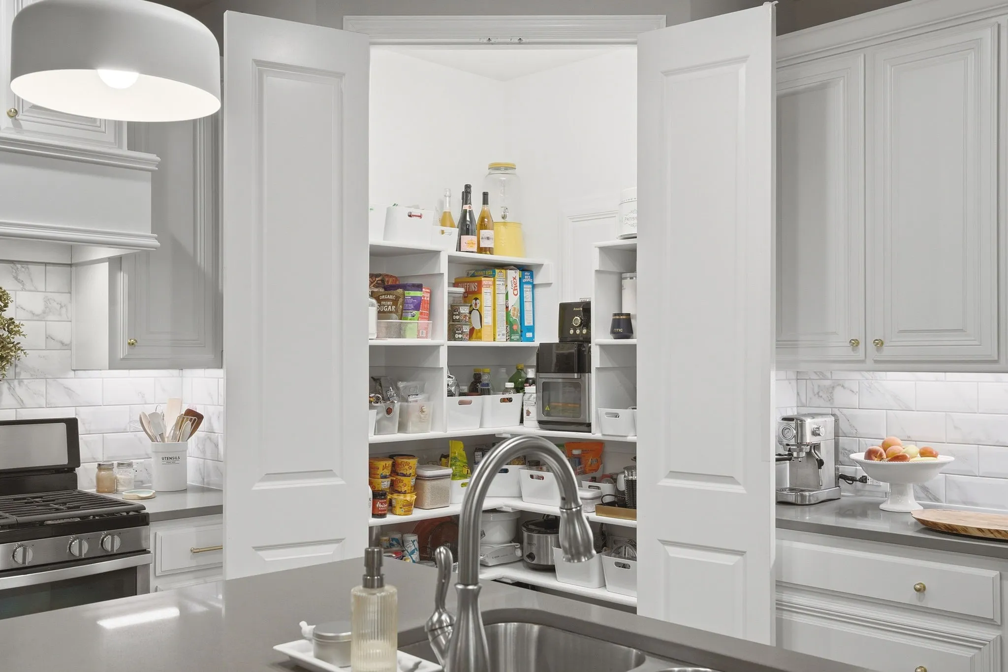 Walk-in pantry with extensive built-in shelving, kitchen island with integrated sink, stainless steel gas range, white subway tile backsplash, and light gray cabinetry with gold-tone hardware