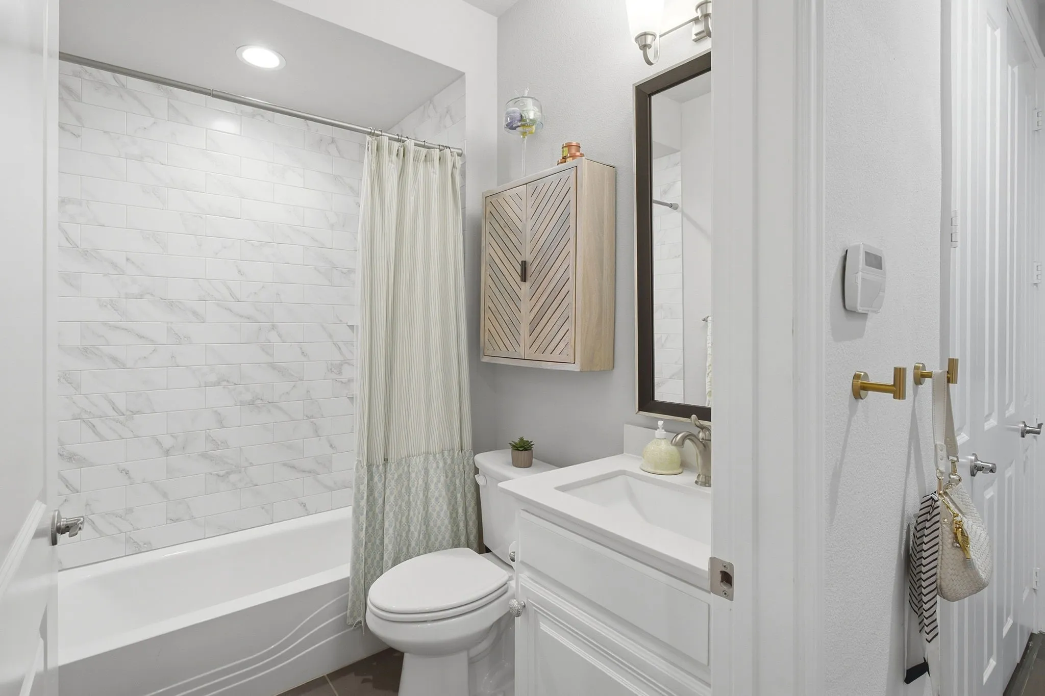 White vanity with integrated sink, rectangular framed mirror, light wood-finish wall cabinet, and tub with tiled surround