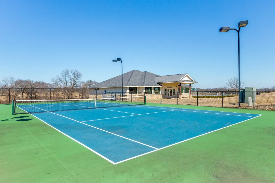 Outdoor tennis court featuring a blue playing surface, green surrounding area, and perimeter fencing