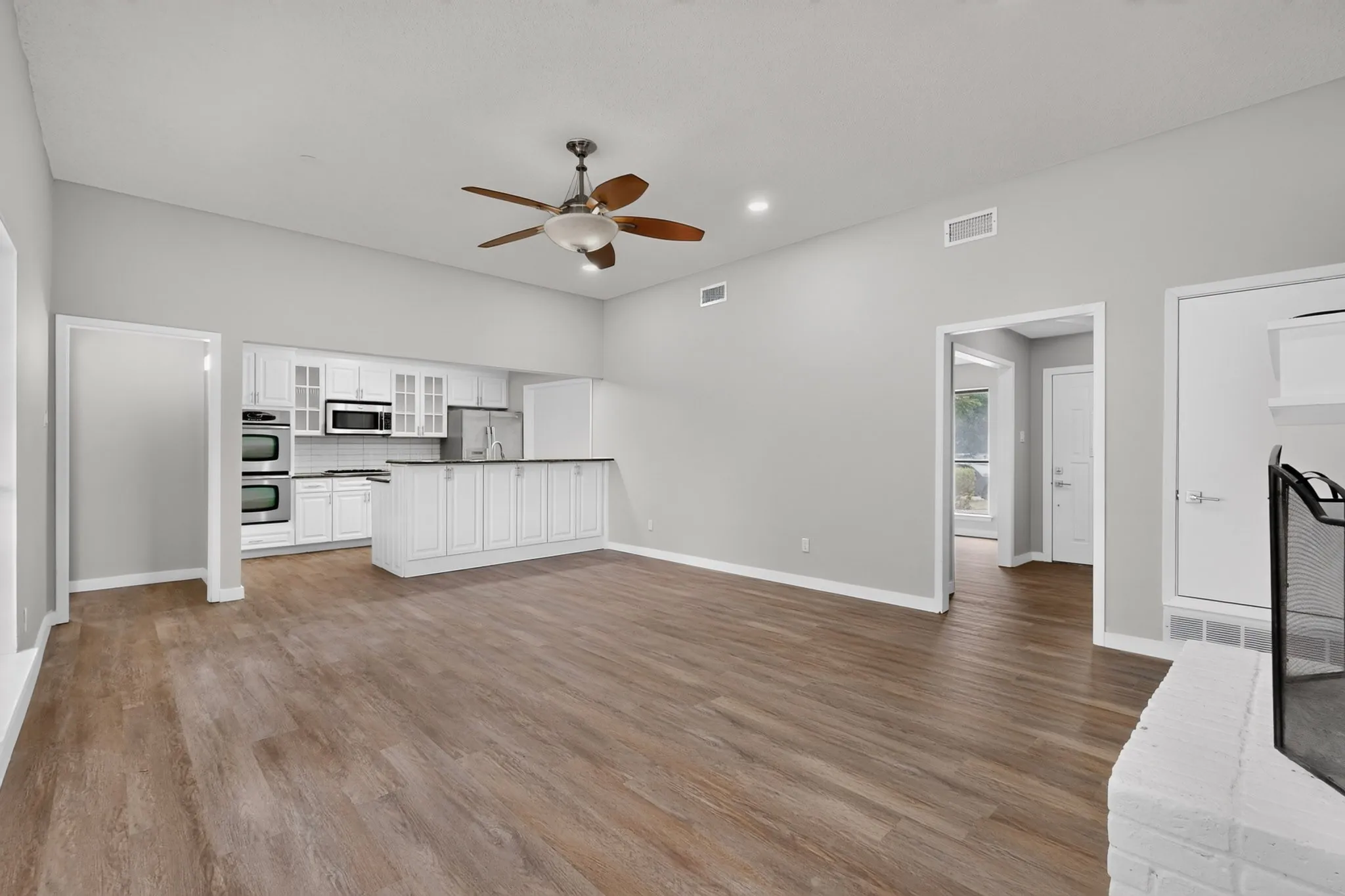 Vaulted ceiling with a ceiling fan, wood-finish flooring, white cabinetry, double wall oven, and a white brick fireplace