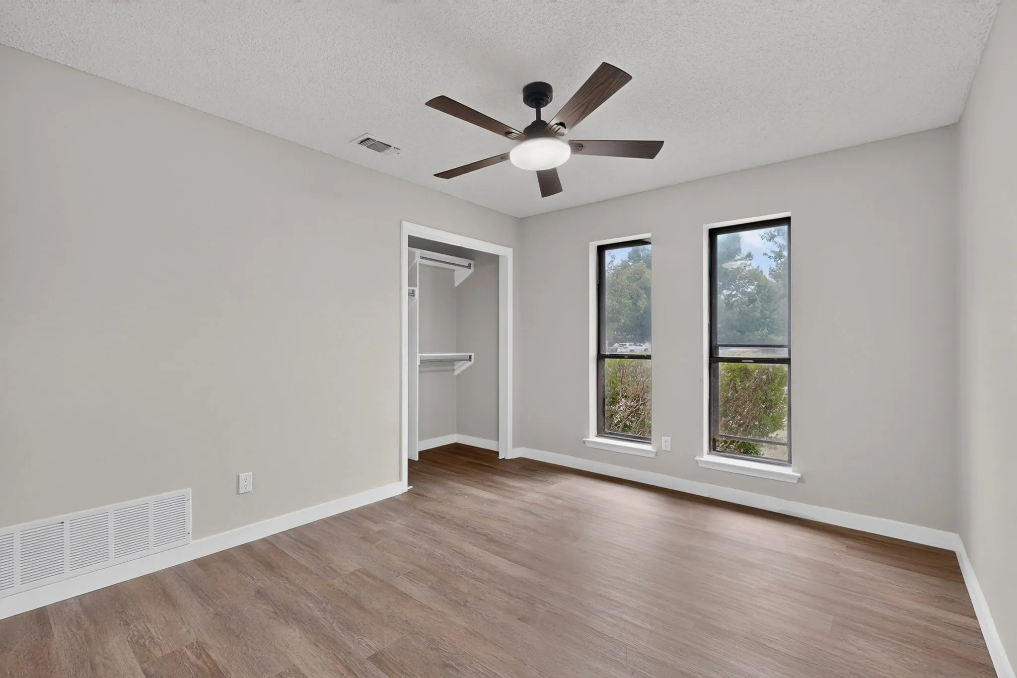 Wood-finish flooring, two large windows, a built-in closet with shelving, a ceiling fan with integrated lighting, and neutral wall tones