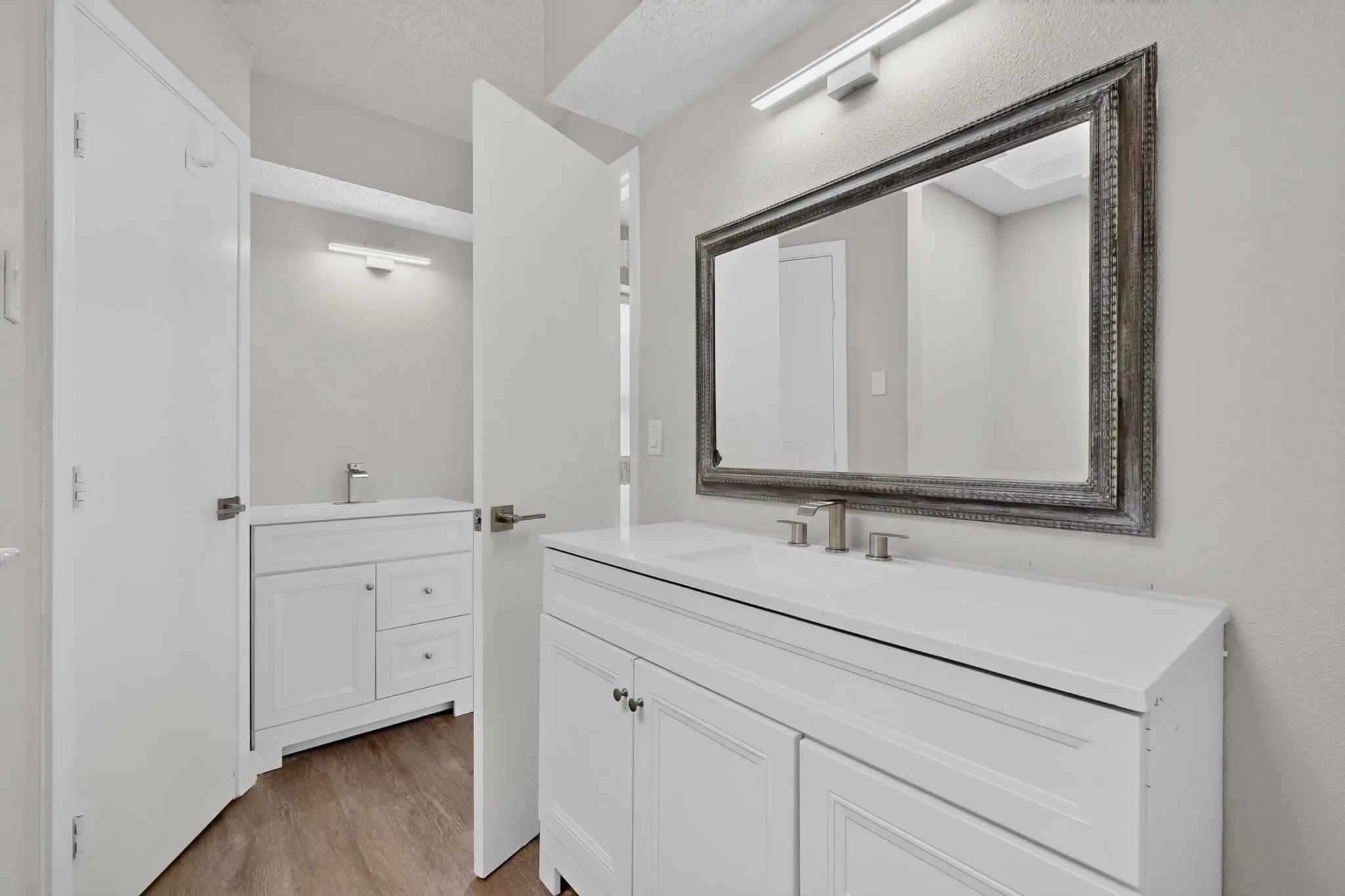 Dual white vanities with integrated sinks, brushed nickel fixtures, custom framed mirror, and wood-finish flooring