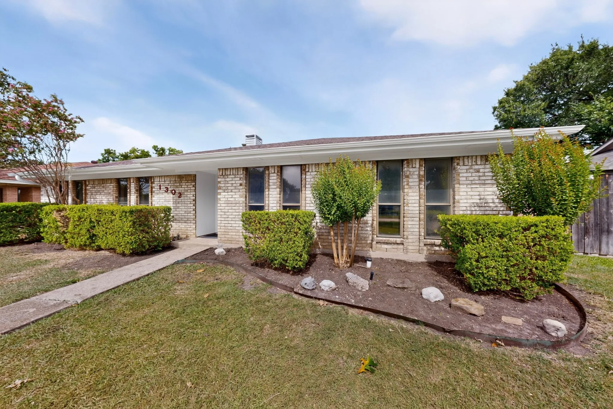 Brick exterior with white trim, featuring a manicured lawn, established landscaping, and a concrete walkway