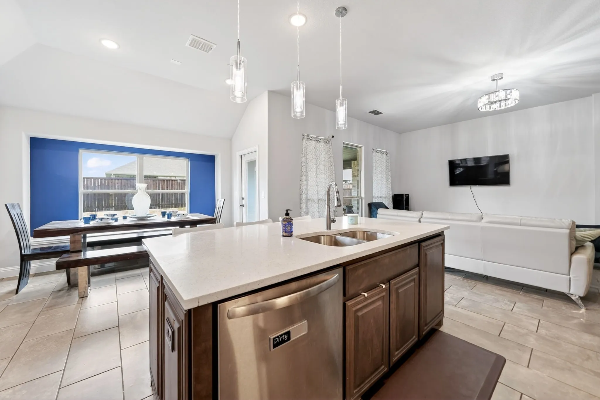 Spacious open-concept layout with vaulted ceilings, a kitchen island featuring a dual basin sink and stainless steel dishwasher, and neutral-toned tile flooring throughout