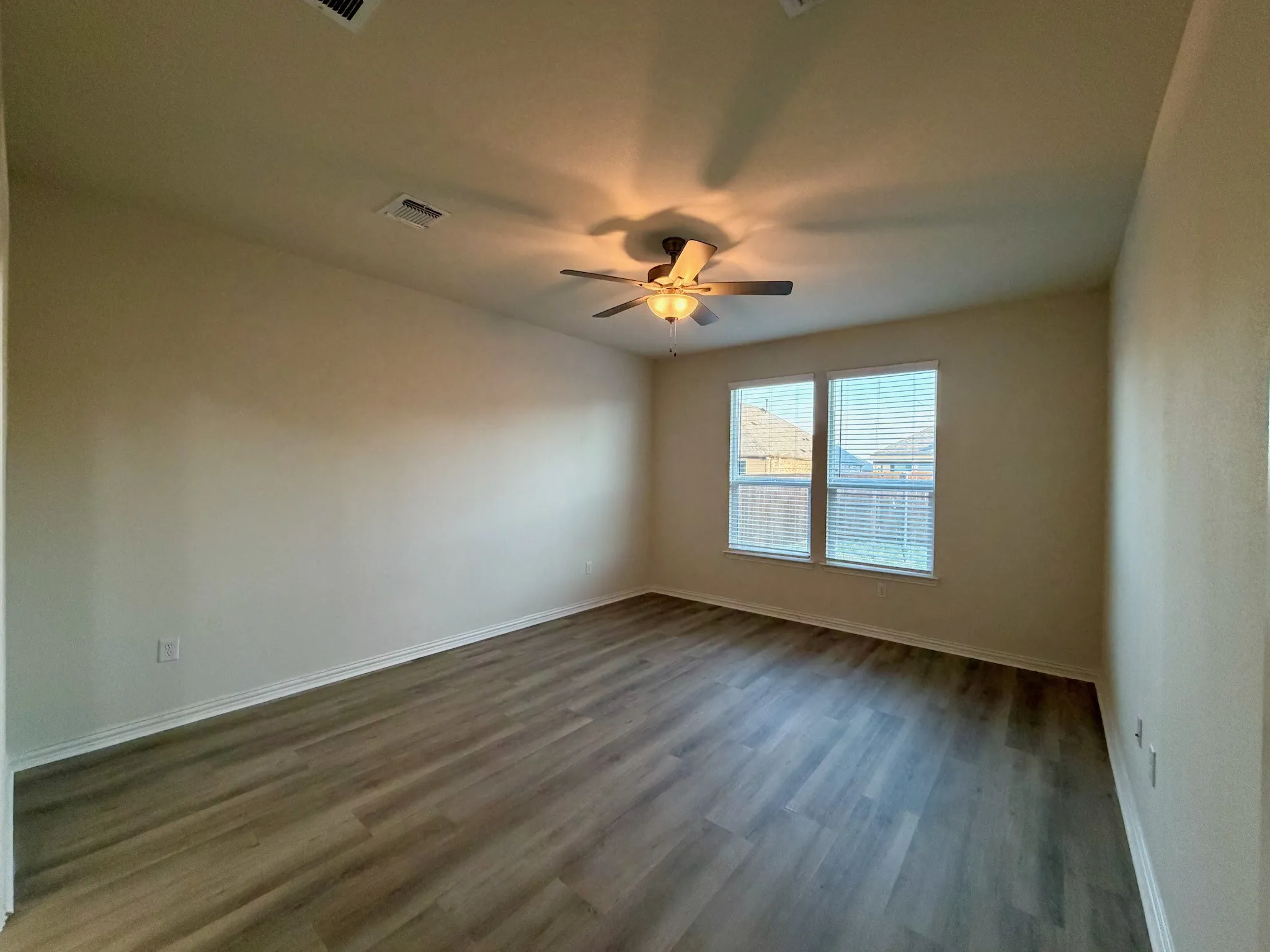 Spacious room with wood-finish flooring, neutral-toned walls, a ceiling fan with integrated lighting, and two windows with horizontal blinds
