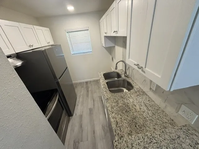 White shaker-style cabinetry, granite countertops, stainless steel double basin sink, chrome gooseneck faucet, and wood-finish flooring