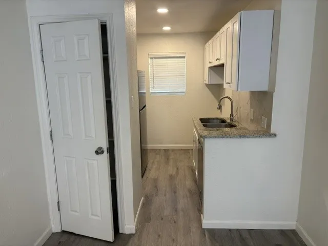 Compact kitchen featuring white upper cabinetry, a double basin sink with a high-arch faucet, a speckled countertop, and wood-finish flooring