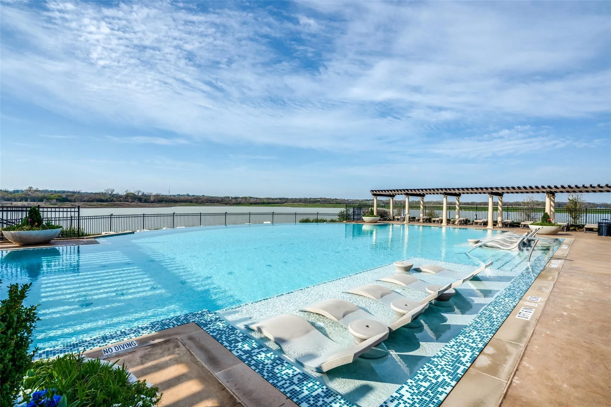 Infinity-edge pool with underwater loungers, integrated spa, and a poolside pergola structure, all overlooking a substantial body of water