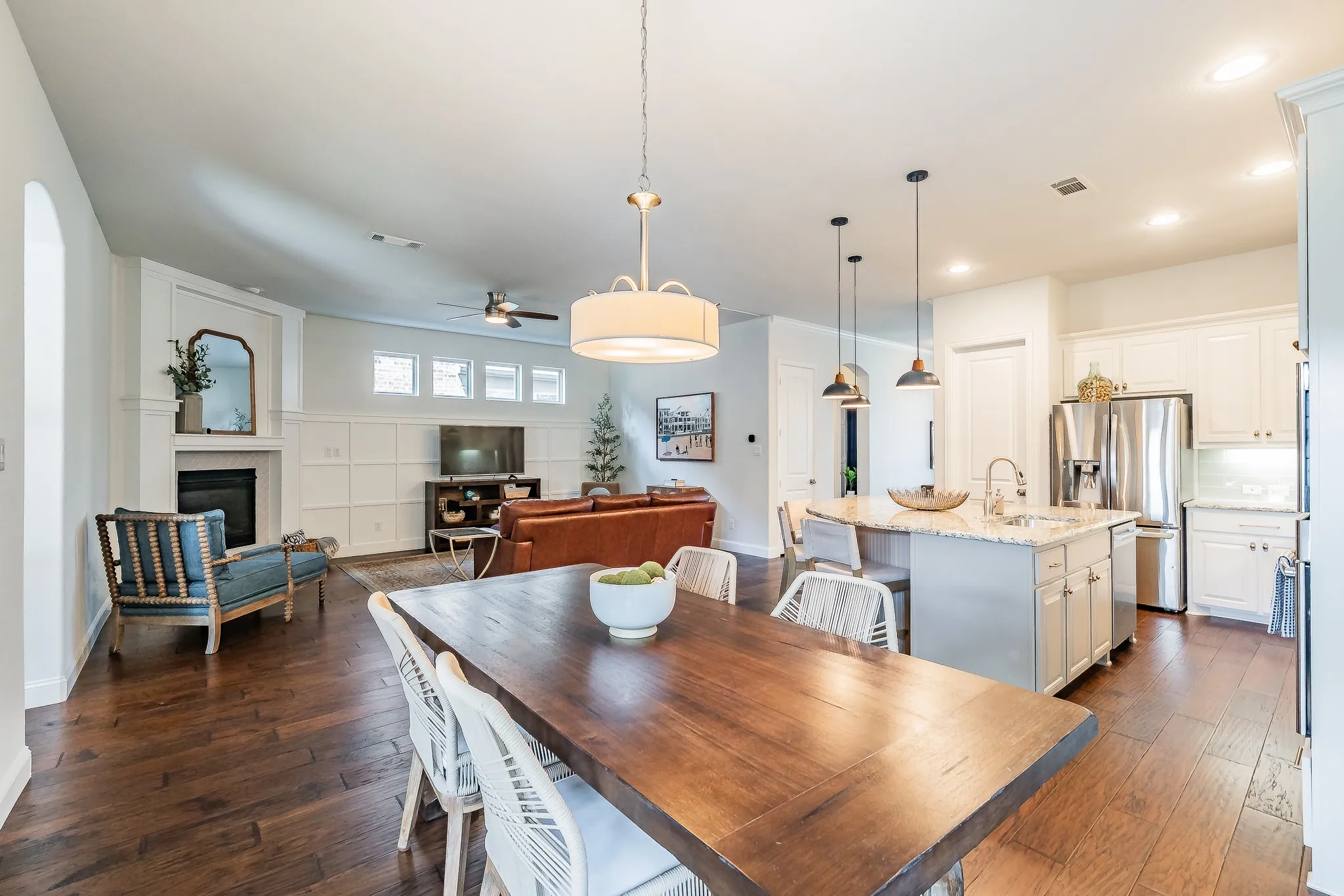 Hardwood-finish flooring throughout, living area with fireplace and wainscoting, kitchen featuring stainless steel appliances, white cabinetry, and a large island with sink, dining area with a drum pendant light fixture