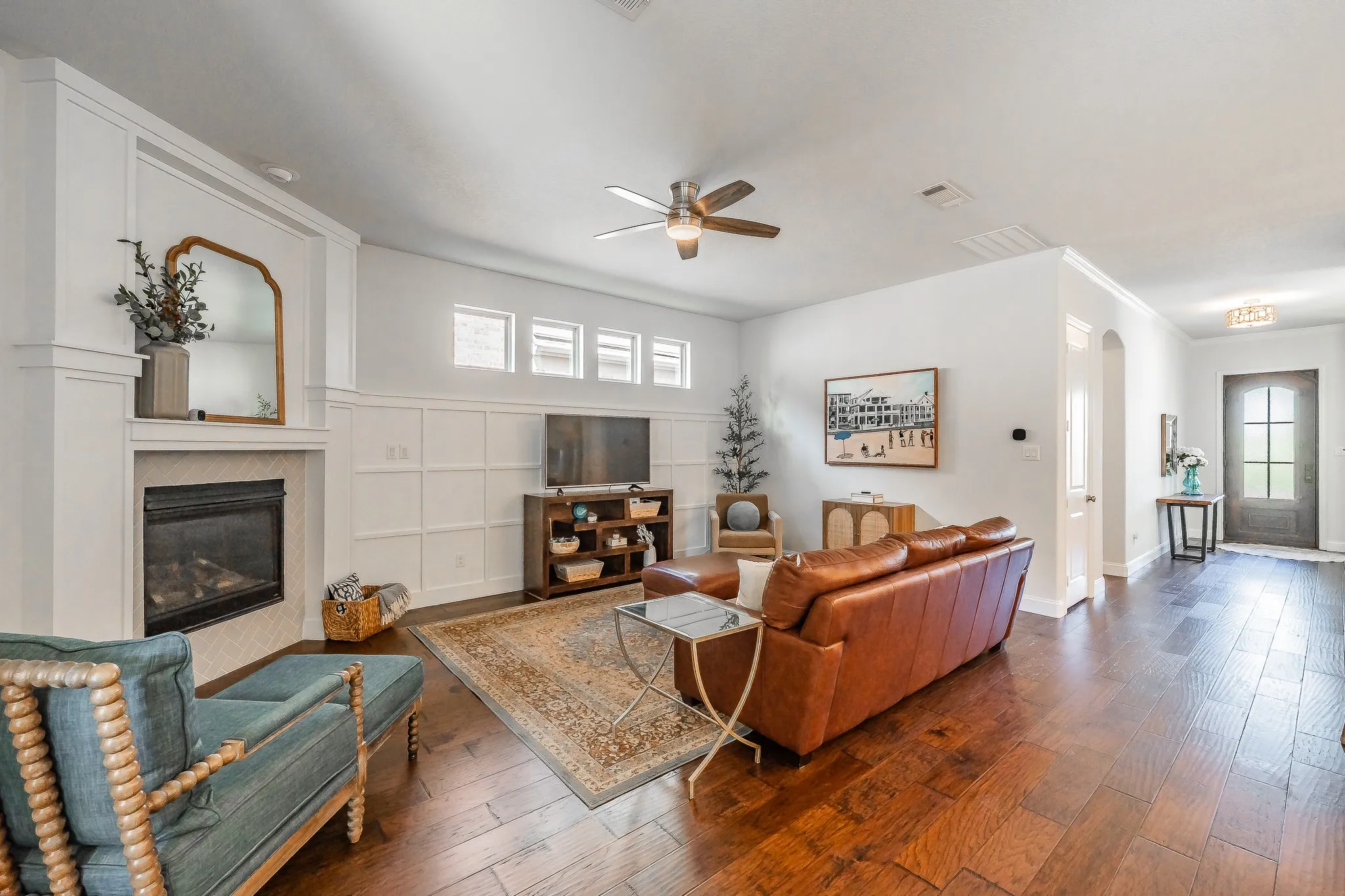 Dark wood-finish flooring extending into the entry, a corner fireplace with white surround, decorative wall paneling, and a ceiling fan with integrated lighting