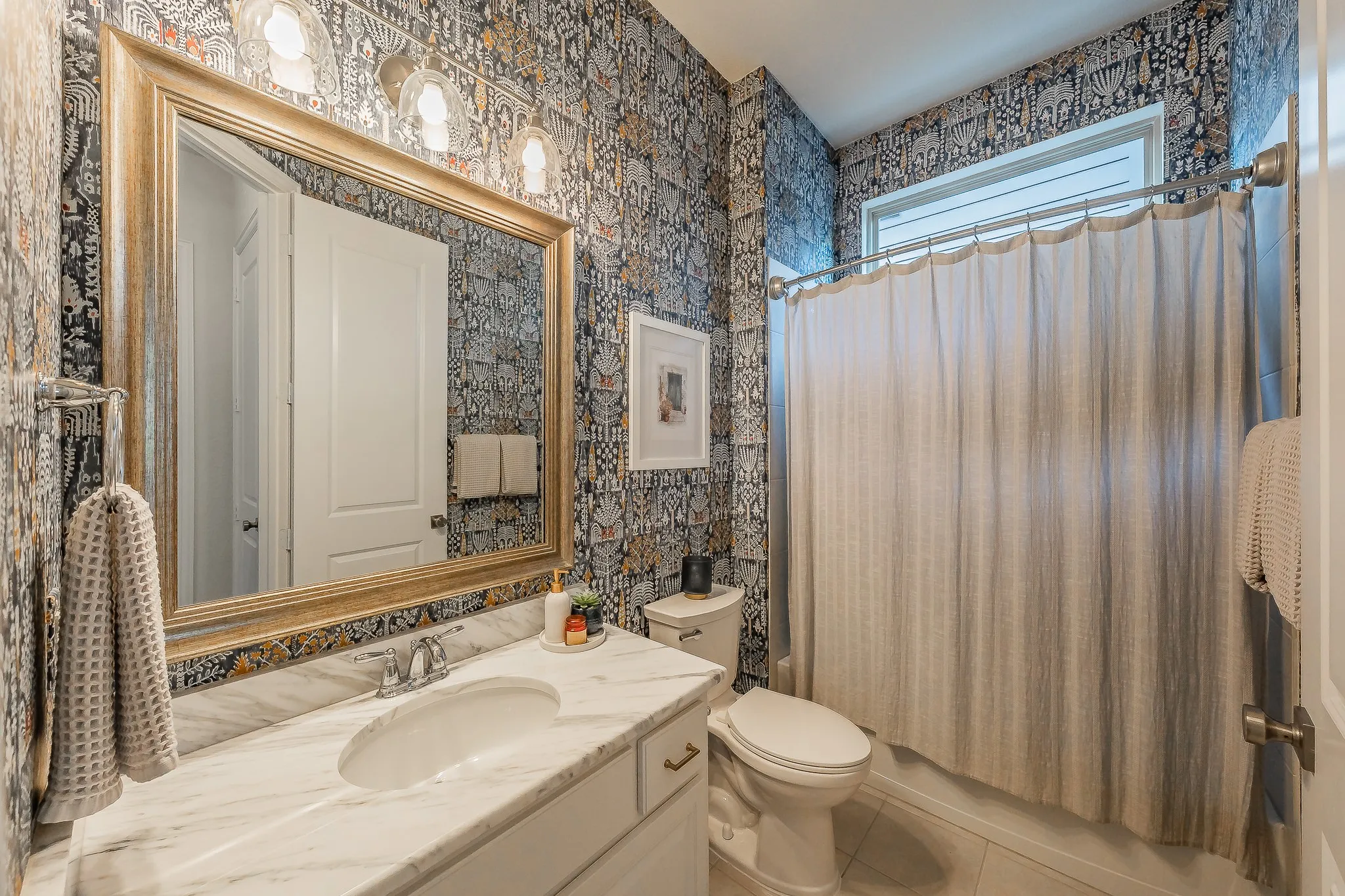 Bathroom featuring patterned wallpaper, a white vanity with an integrated sink, a large framed mirror, and a window with a shower curtain