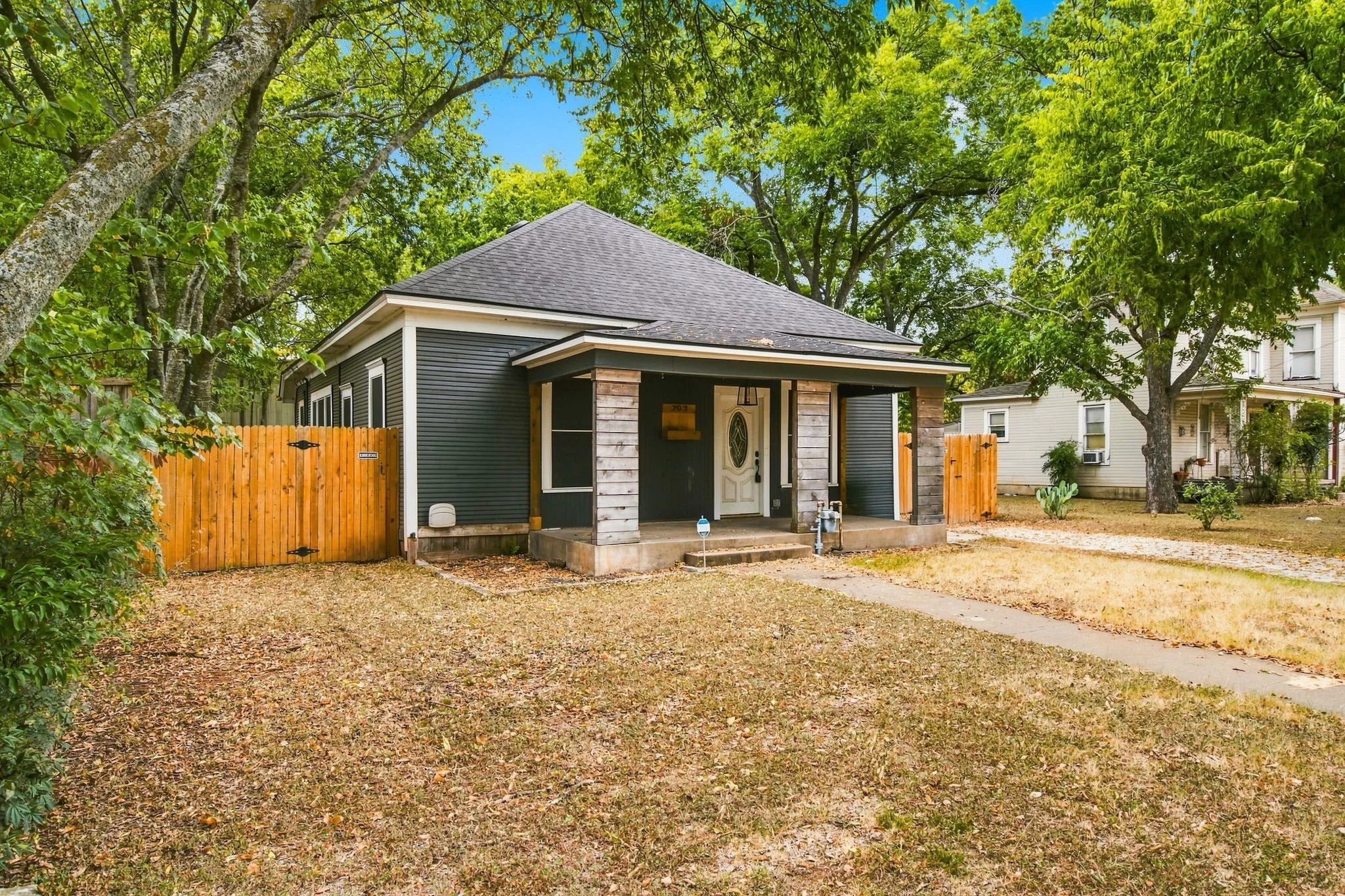 Dark-toned exterior siding, covered front porch with stone-clad columns, white paneled front door, and a wooden privacy fence