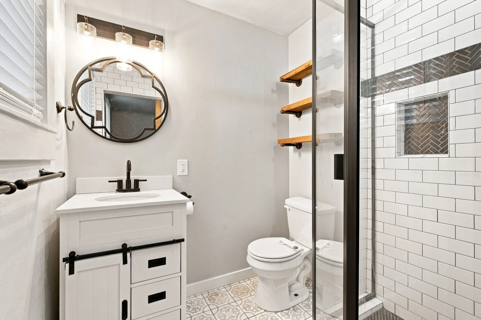 White vanity with barn door accent and matte black fixtures, decorative floor tiling, walk-in shower with subway tile and herringbone accent, integrated shower niche, and glass enclosure