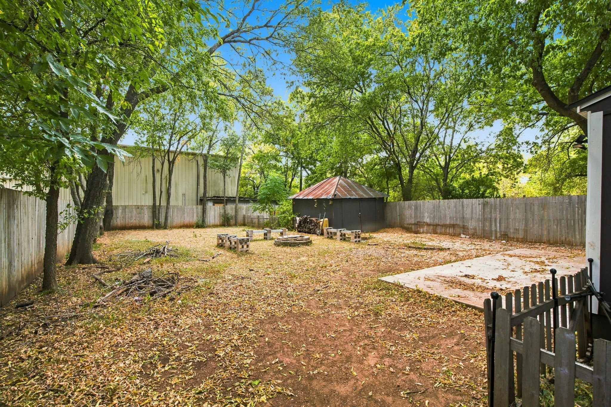 Expansive yard featuring a wooden privacy fence, mature trees, a concrete patio, and an accessory building with a metal roof