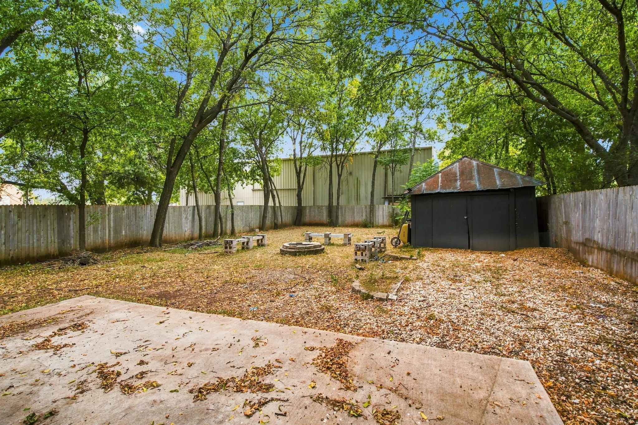 Spacious backyard featuring a concrete patio, mature trees, wood privacy fencing, a fire pit area, and a storage shed with a corrugated metal roof