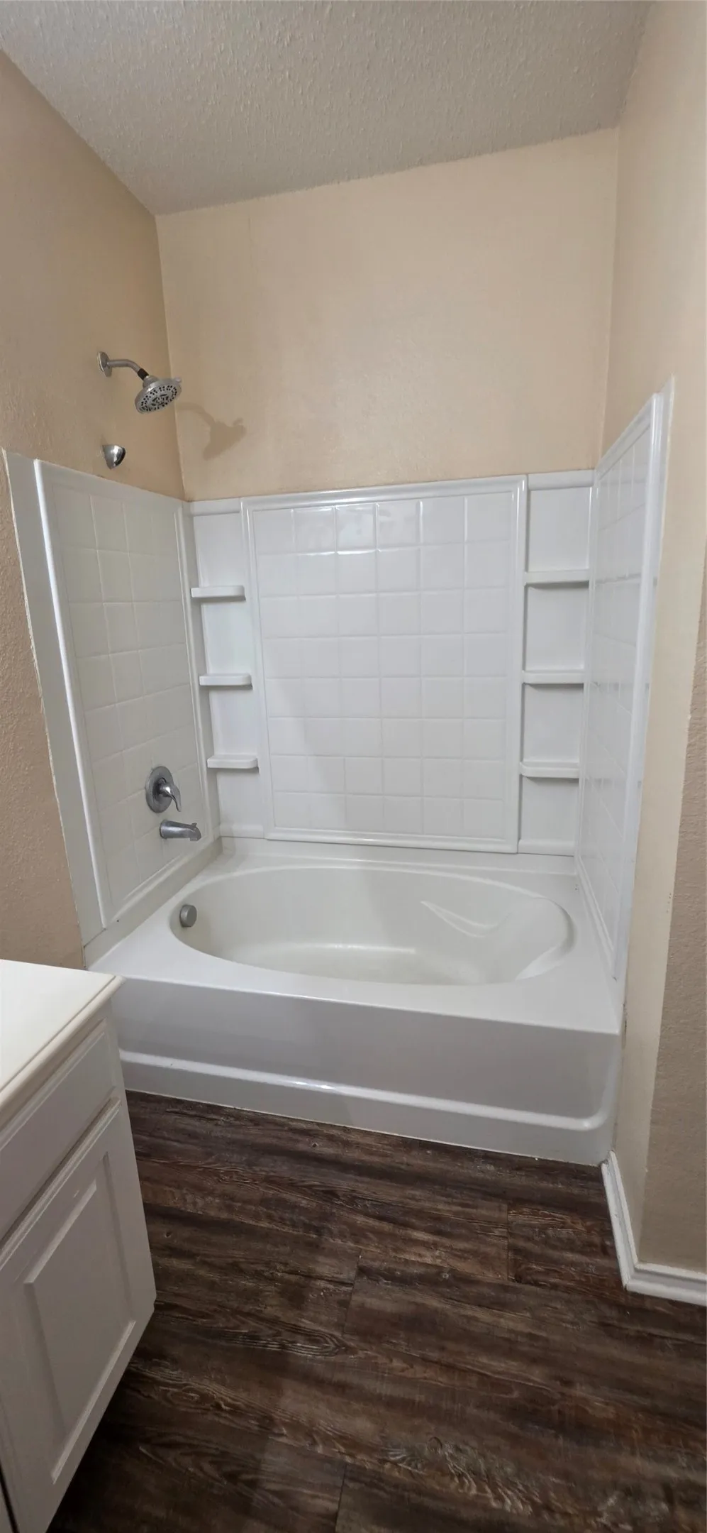 Bathtub with integrated shower surround and built-in shelving, chrome fixtures, and wood-finish flooring