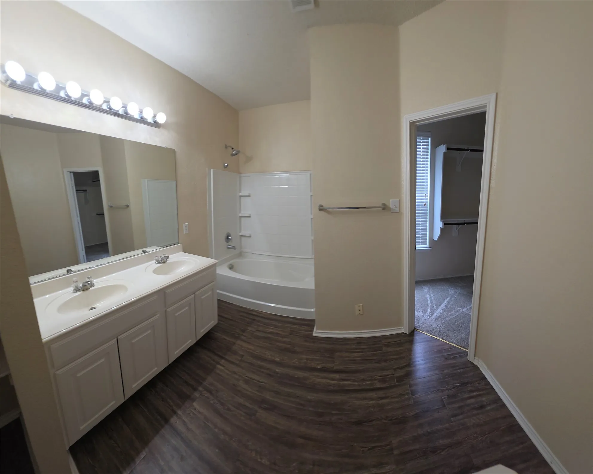 Dual vanity with integrated sinks, wood-finish flooring, built-in bathtub with shower, and an adjacent room with a window