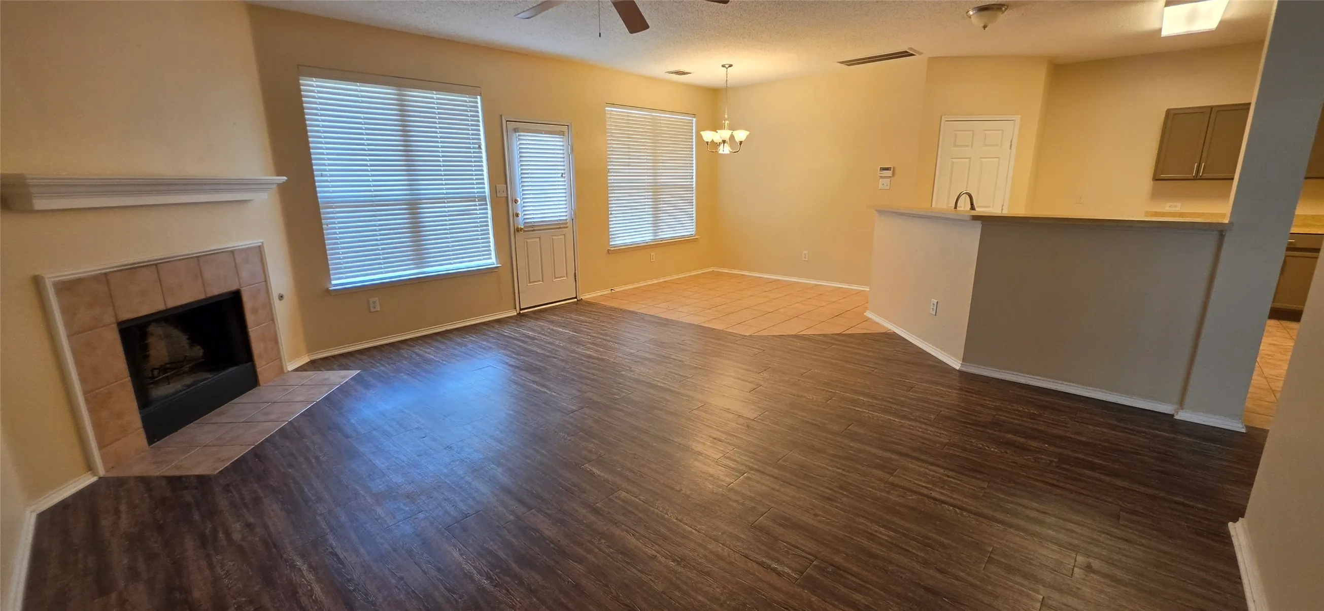 Wood-finish flooring, tiled fireplace, multiple windows with blinds, built-in breakfast bar, and a ceiling fan with integrated lighting
