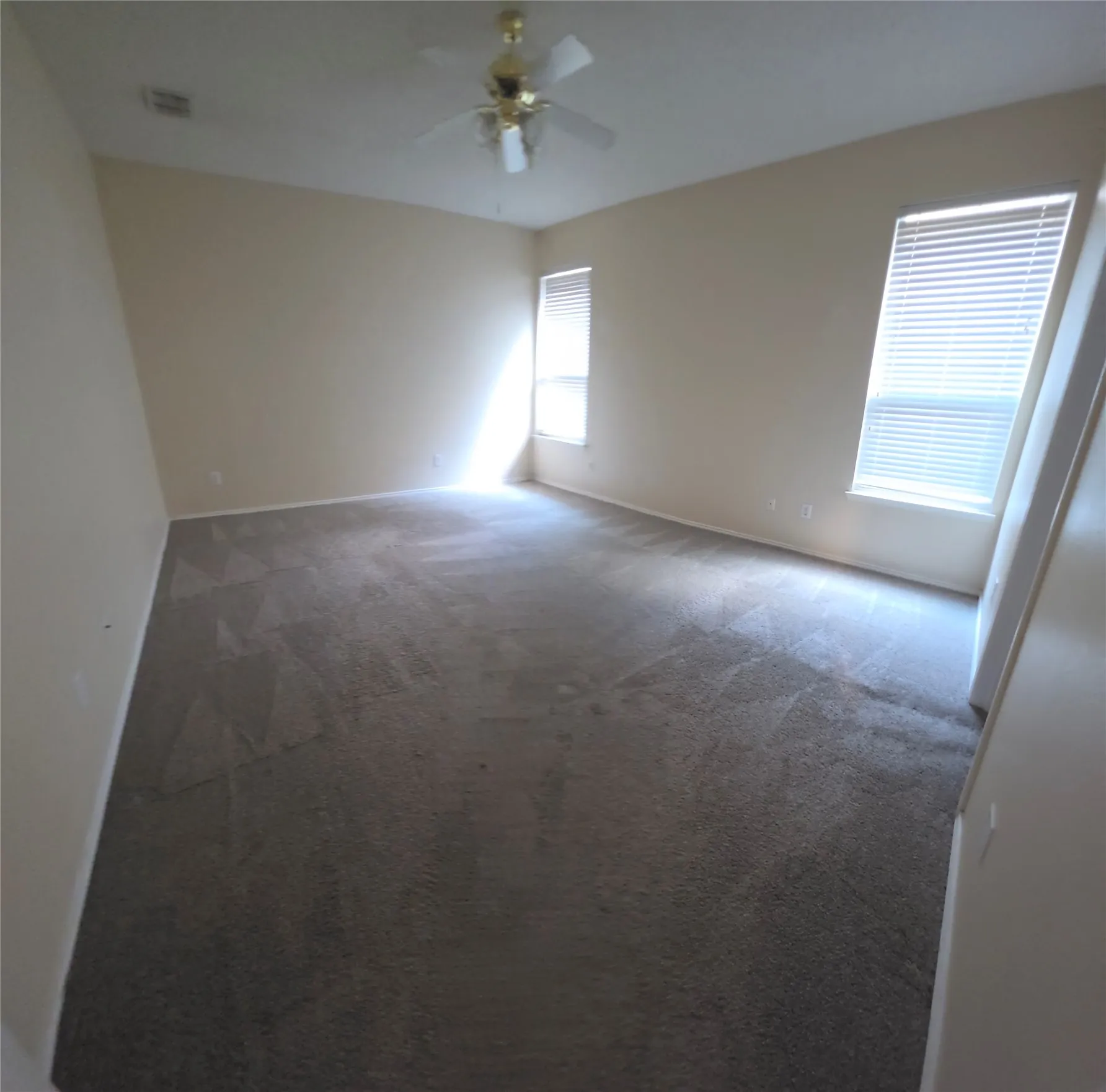 Warm neutral wall paint, ceiling fan with light fixture, two windows with blinds, and dark gray carpeting