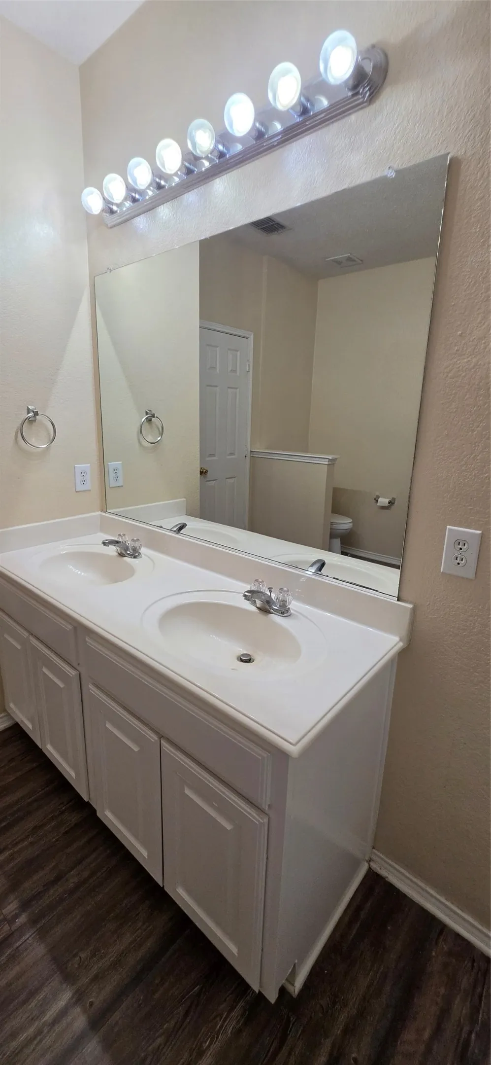 Dual vanity with integrated sinks, white cabinetry, dark wood-finish flooring, and an expansive wall mirror with overhead lighting