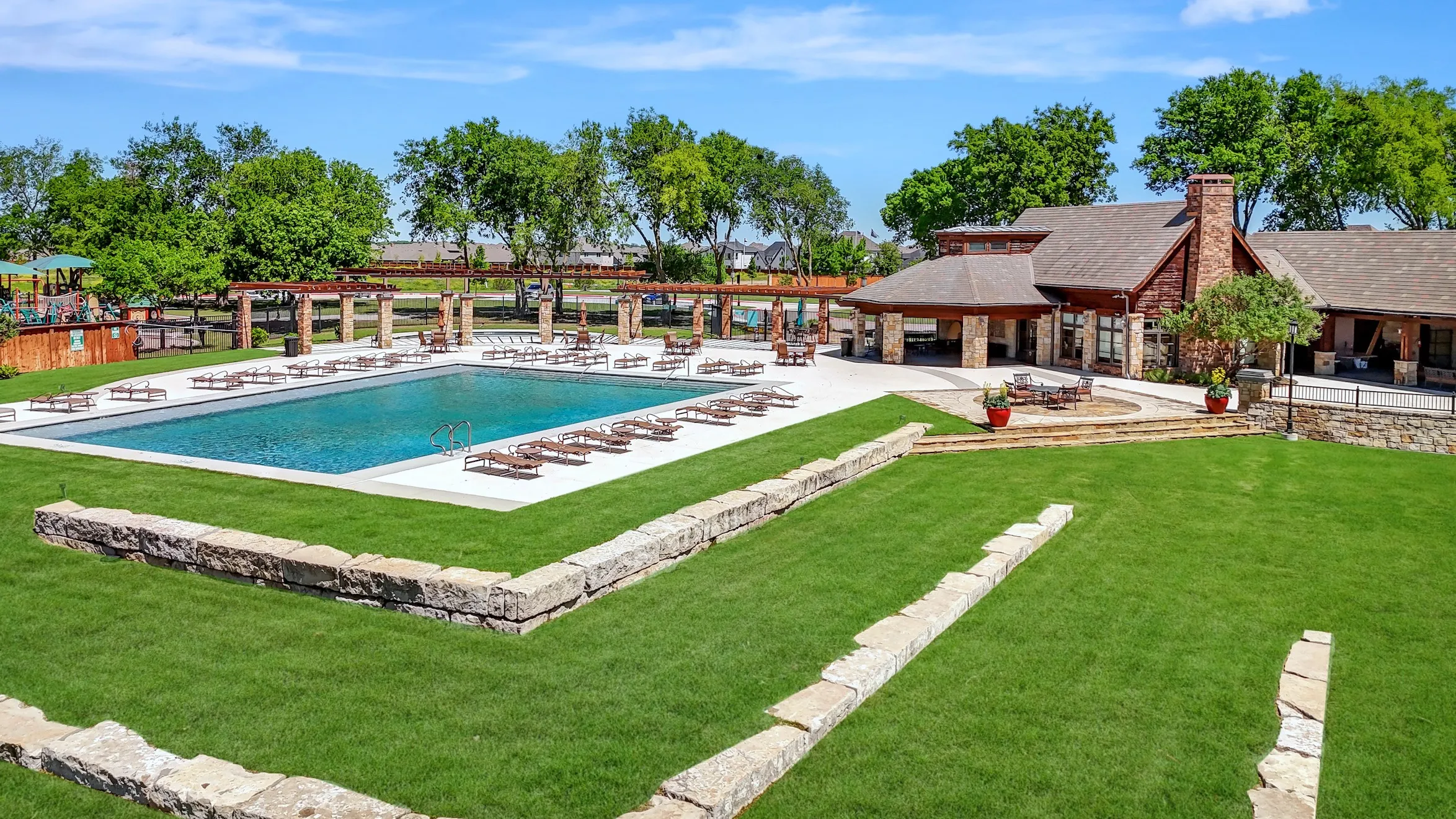 Expansive green lawn, stone retaining walls, large rectangular swimming pool with surrounding lounge seating, and a clubhouse building featuring a stone and brick exterior with a prominent chimney