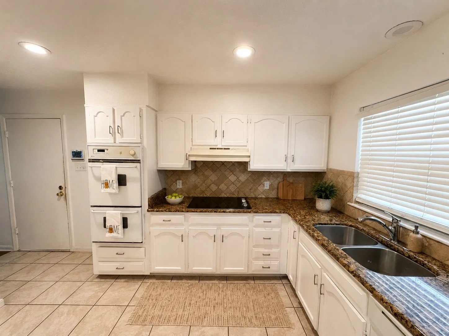 White cabinetry with arched panel doors, stone tile backsplash, granite countertops, double basin stainless steel sink, and tile flooring - virtual staging on countertops