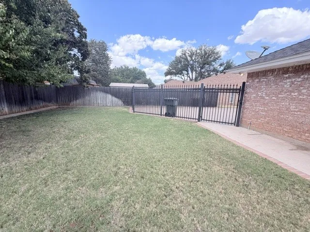 Expansive backyard featuring a privacy fence, wrought iron gate, brick exterior, and concrete pathway with brick edging