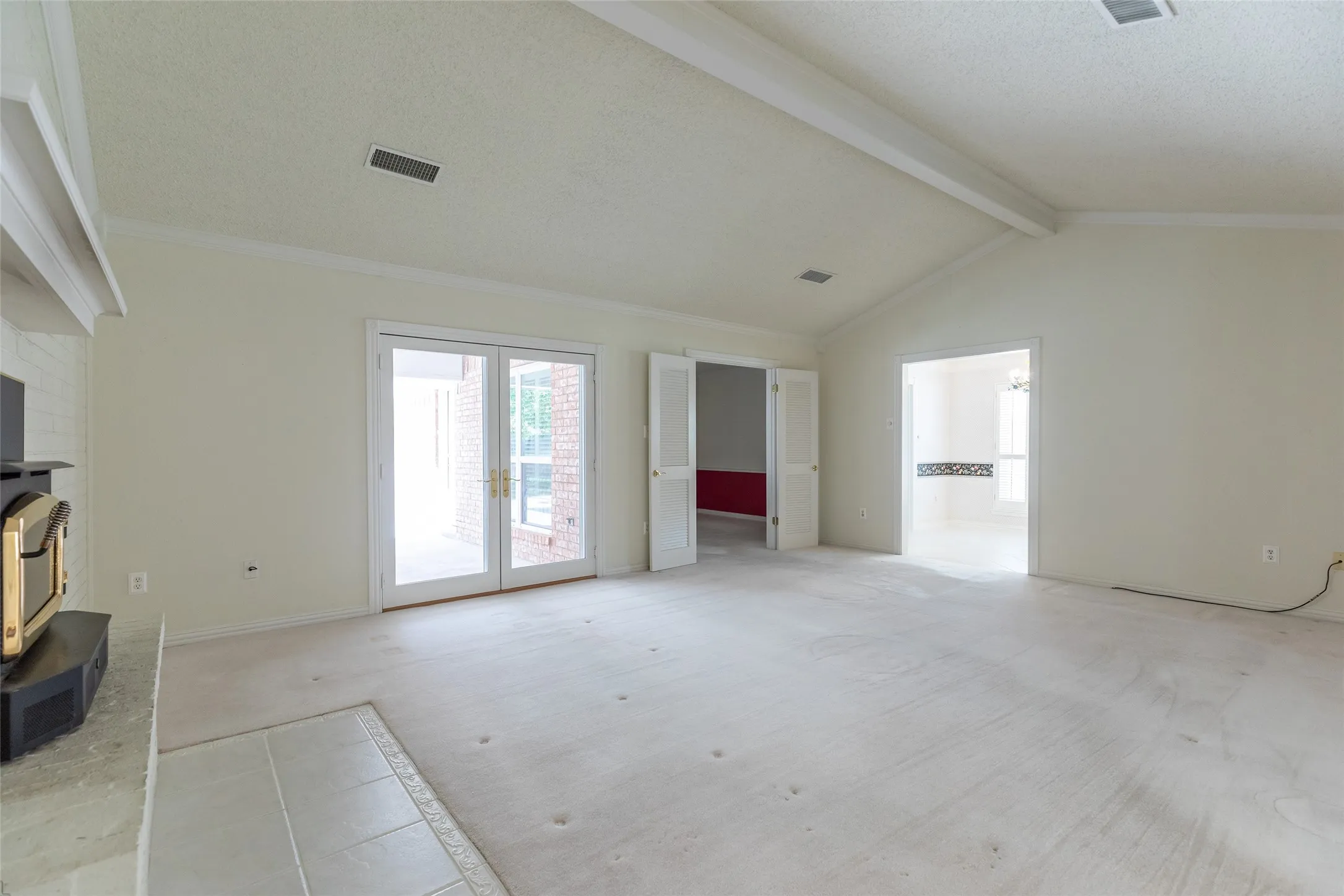 Vaulted ceiling with exposed beams, double glass-paneled French doors, fireplace with tiled hearth, and crown molding