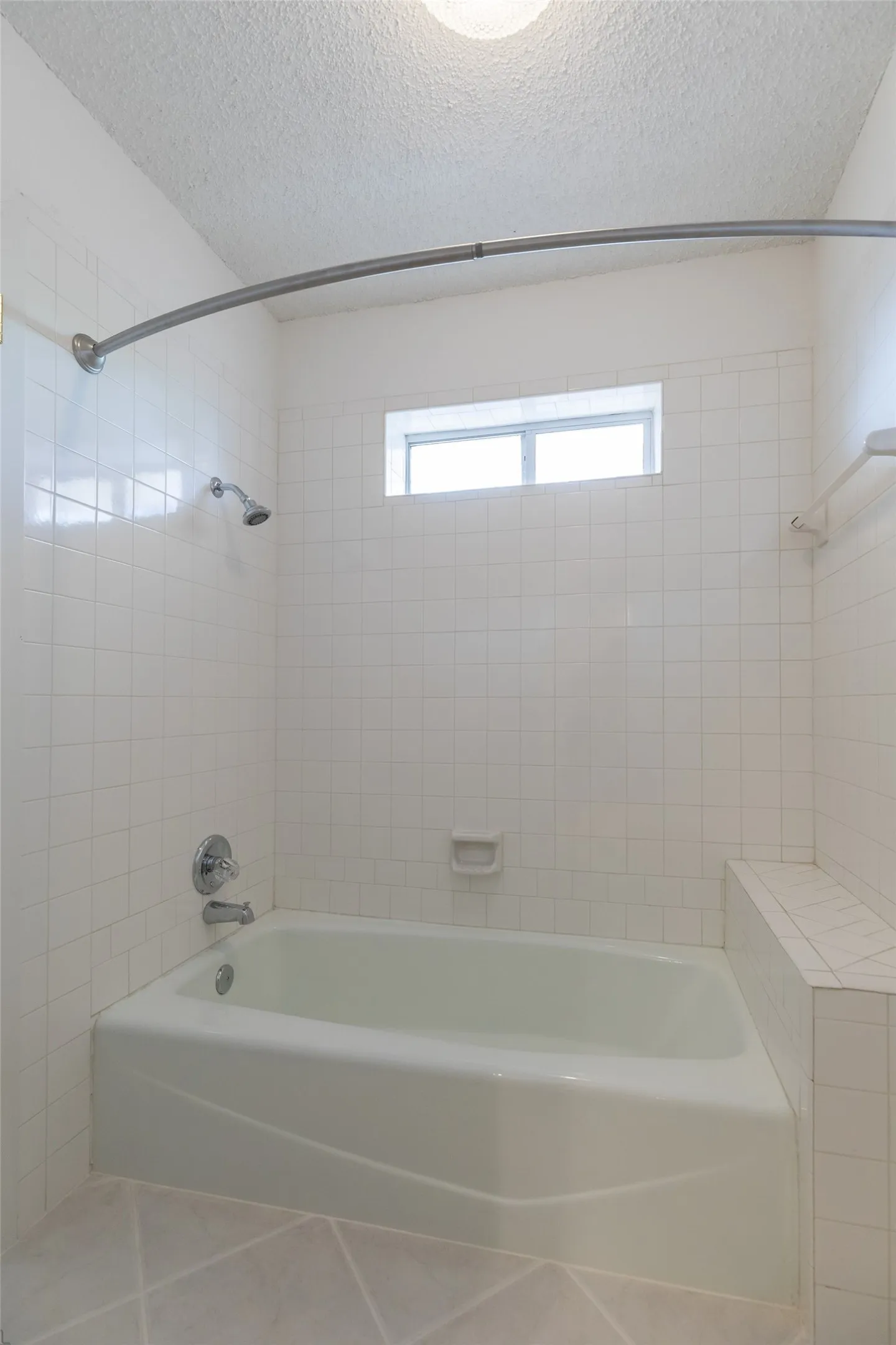 Bathtub with white tile surround, integrated shower, and a high-set window