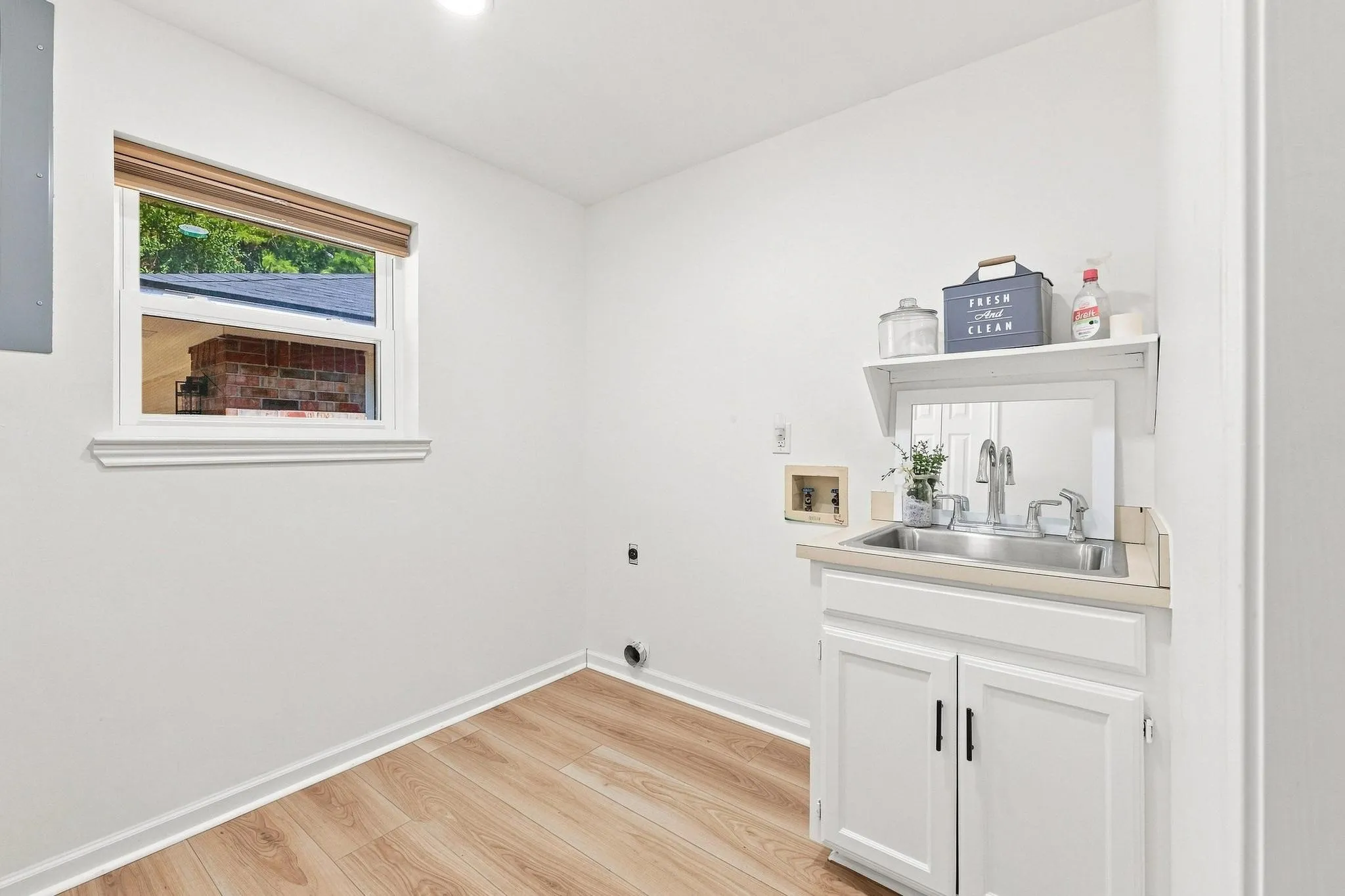Utility sink with chrome faucet, light wood-finish flooring, white wall cabinetry, and a window with a neutral-toned blind