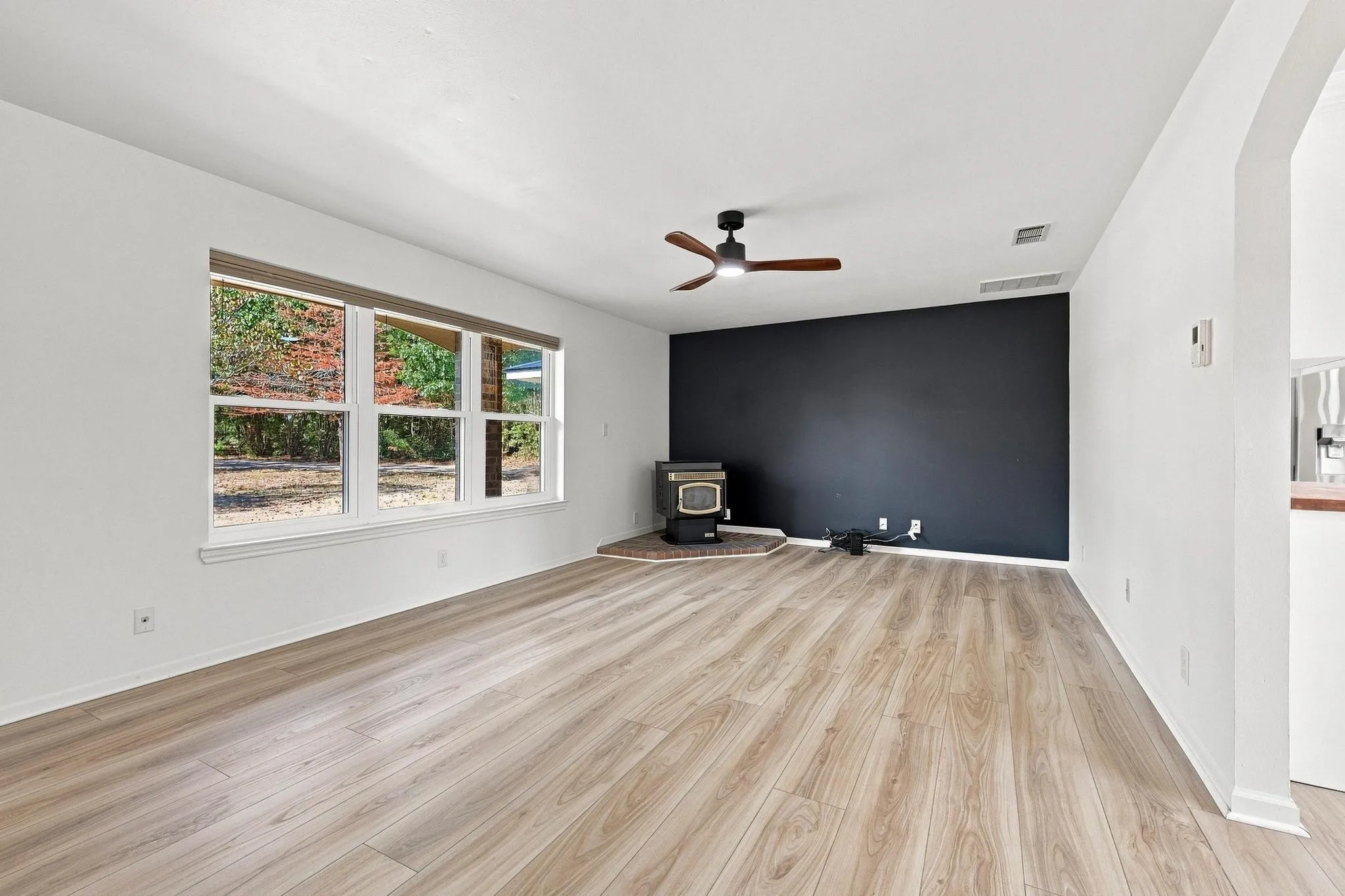 Bright, open living space showcasing wood-finish flooring throughout, expansive windows with natural light, a dark accent wall, ceiling fan, and clean white walls with architectural detail
