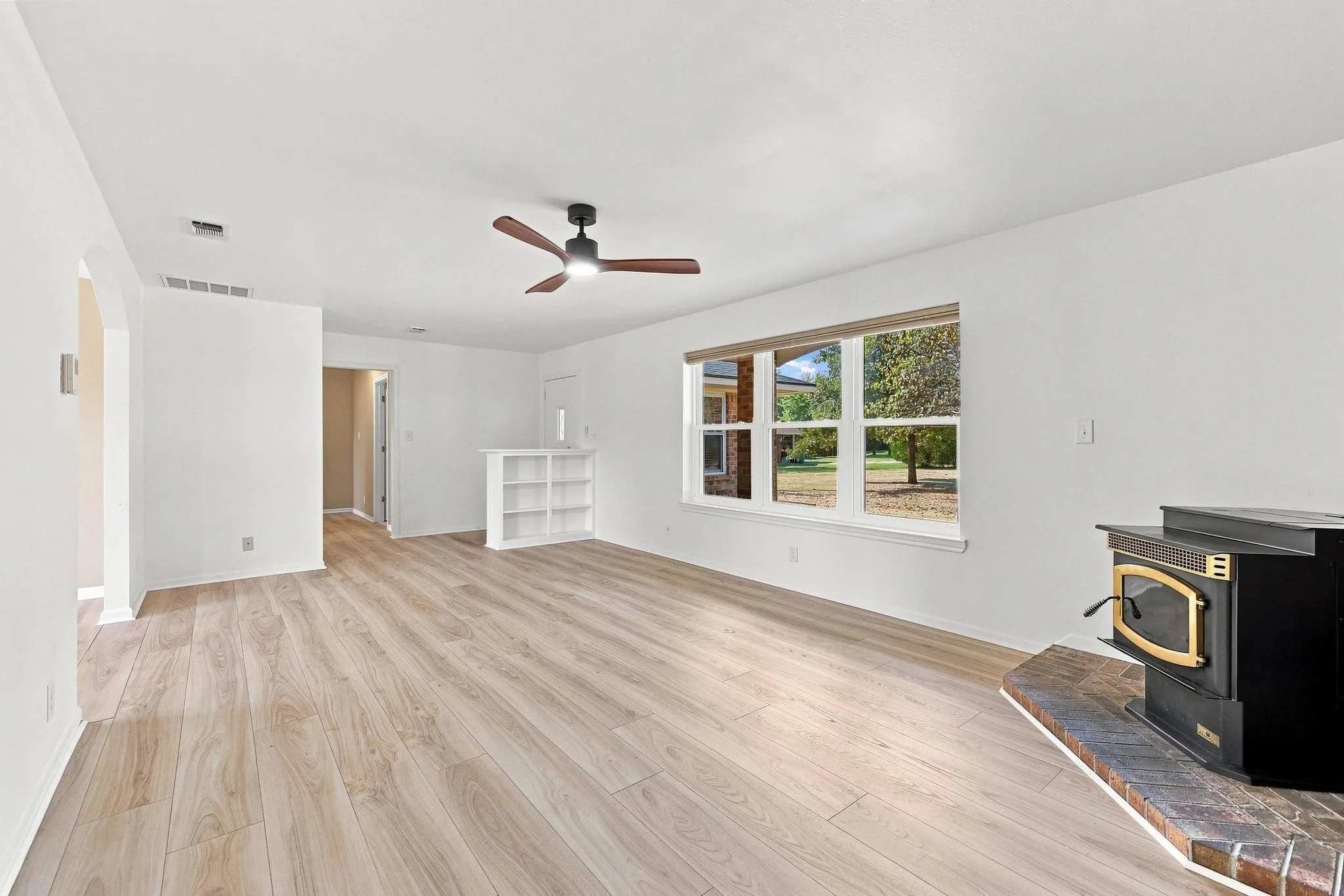 Spacious room featuring wood-finish flooring, a built-in wood stove on a brick hearth, a large multi-pane window, a ceiling fan, and a built-in low bookcase