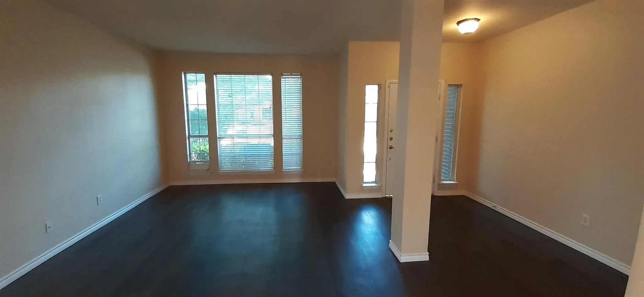 Expansive open-concept room featuring dark wood-finish flooring, multiple windows with blinds, a white entry door with side lights, and a decorative ceiling light fixture