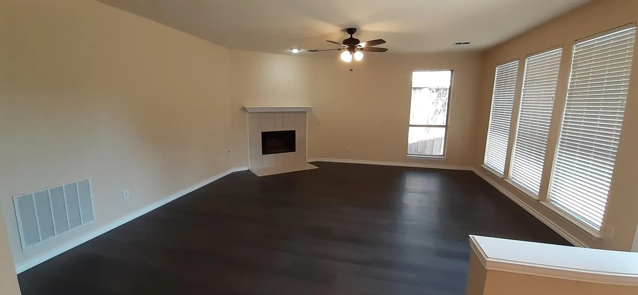 Spacious living area featuring wood-finish flooring, a tiled fireplace with a mantel, multiple windows with blinds, and a ceiling fan with integrated lighting