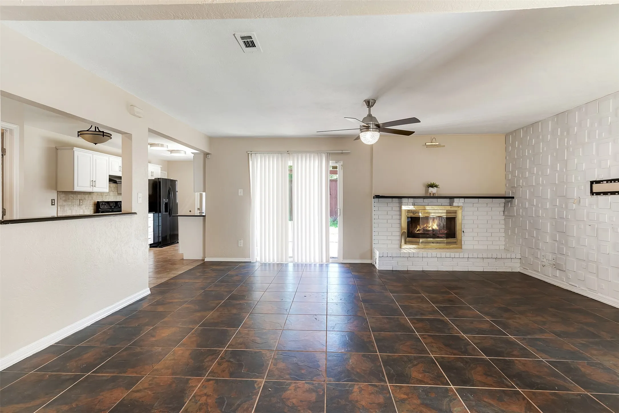 Tile flooring, brick fireplace with a brass-finish insert, white accent wall, ceiling fan, and sliding glass door with vertical blinds