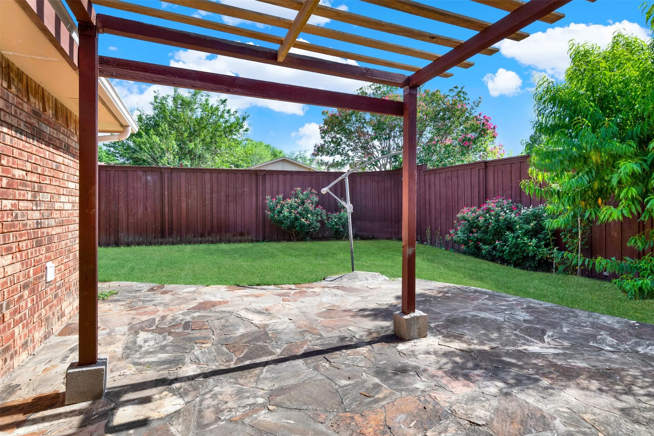 Expansive stone patio with overhead pergola, brick exterior, fenced yard, and established landscaping
