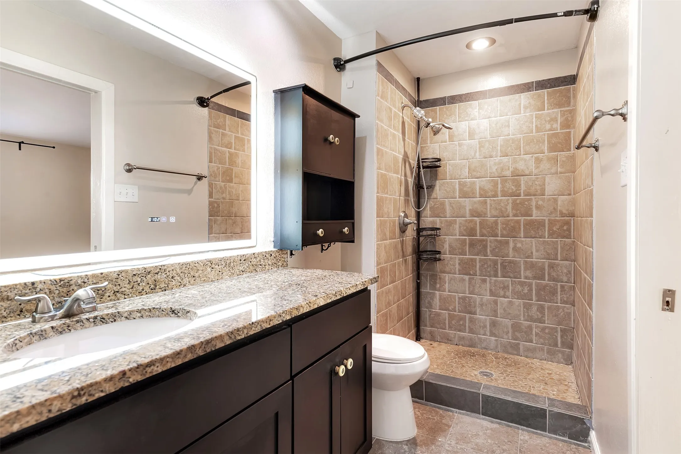 Walk-in shower with natural stone-look tiling, granite-look countertop, dark wood-finish vanity, and illuminated mirror
