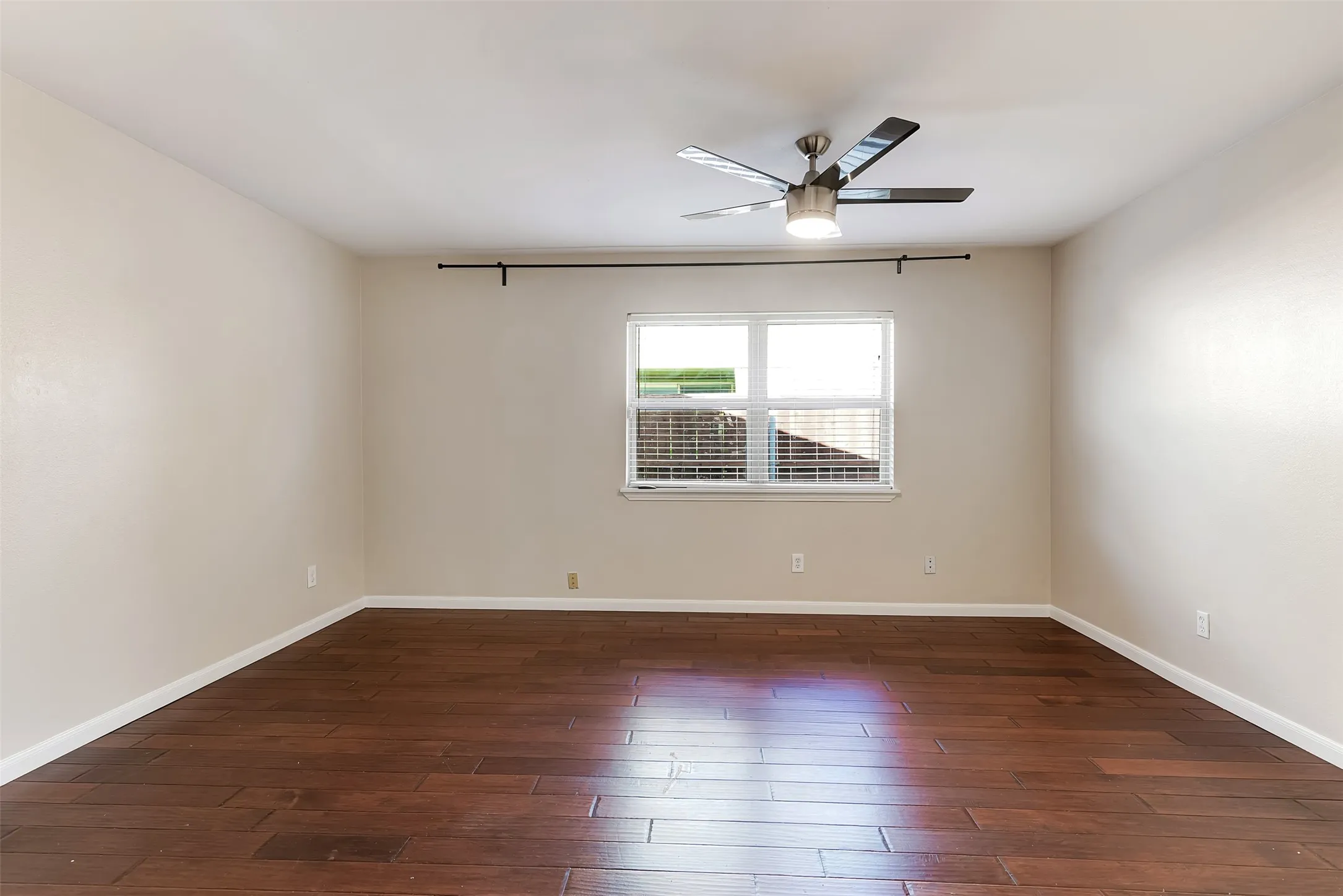 Spacious room with wood-finish flooring, neutral-toned walls, a window with horizontal blinds, and a ceiling fan with integrated lighting