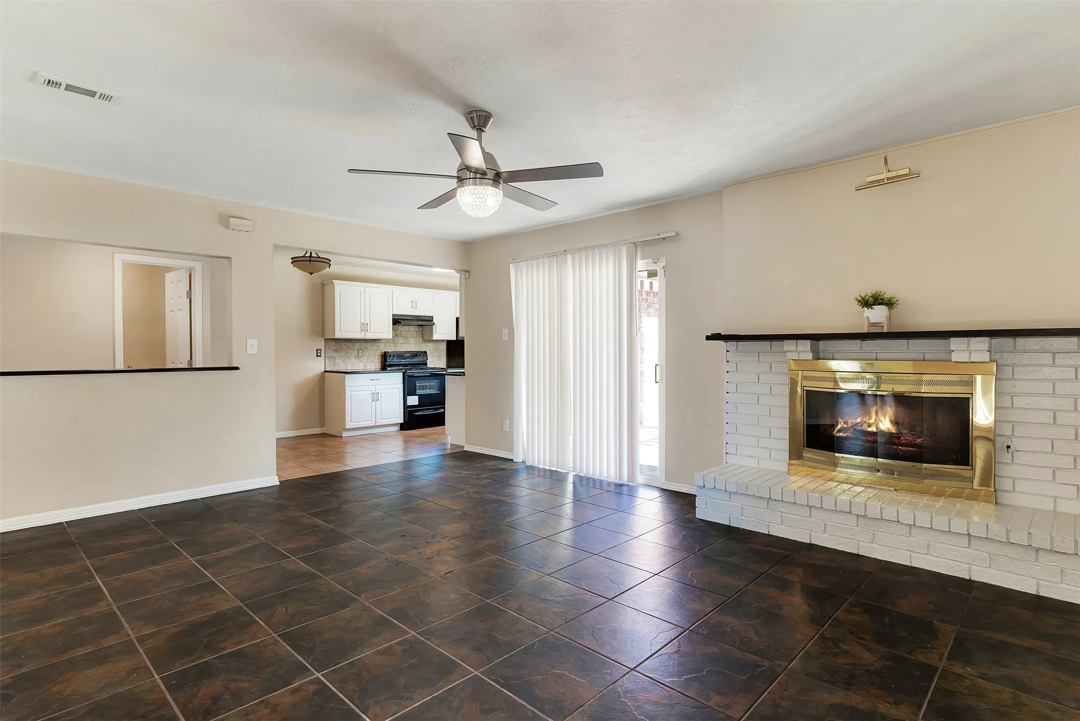 Tile flooring, white brick fireplace with brass trim, sliding glass door, ceiling fan with integrated lighting, and kitchen pass-through window