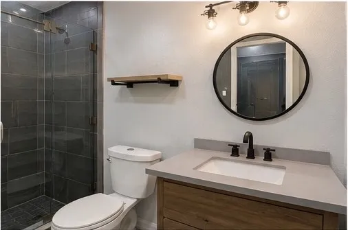 Glass-enclosed shower with dark tile surround, light wood-finish vanity with a single basin sink, light-toned countertop, and matte black fixtures