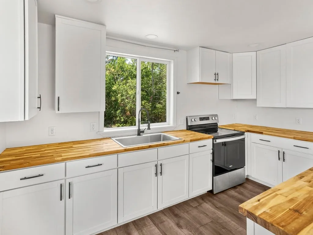 White shaker cabinetry, wood-finish countertops, stainless steel sink, black wall-mounted faucet, and wood-finish flooring